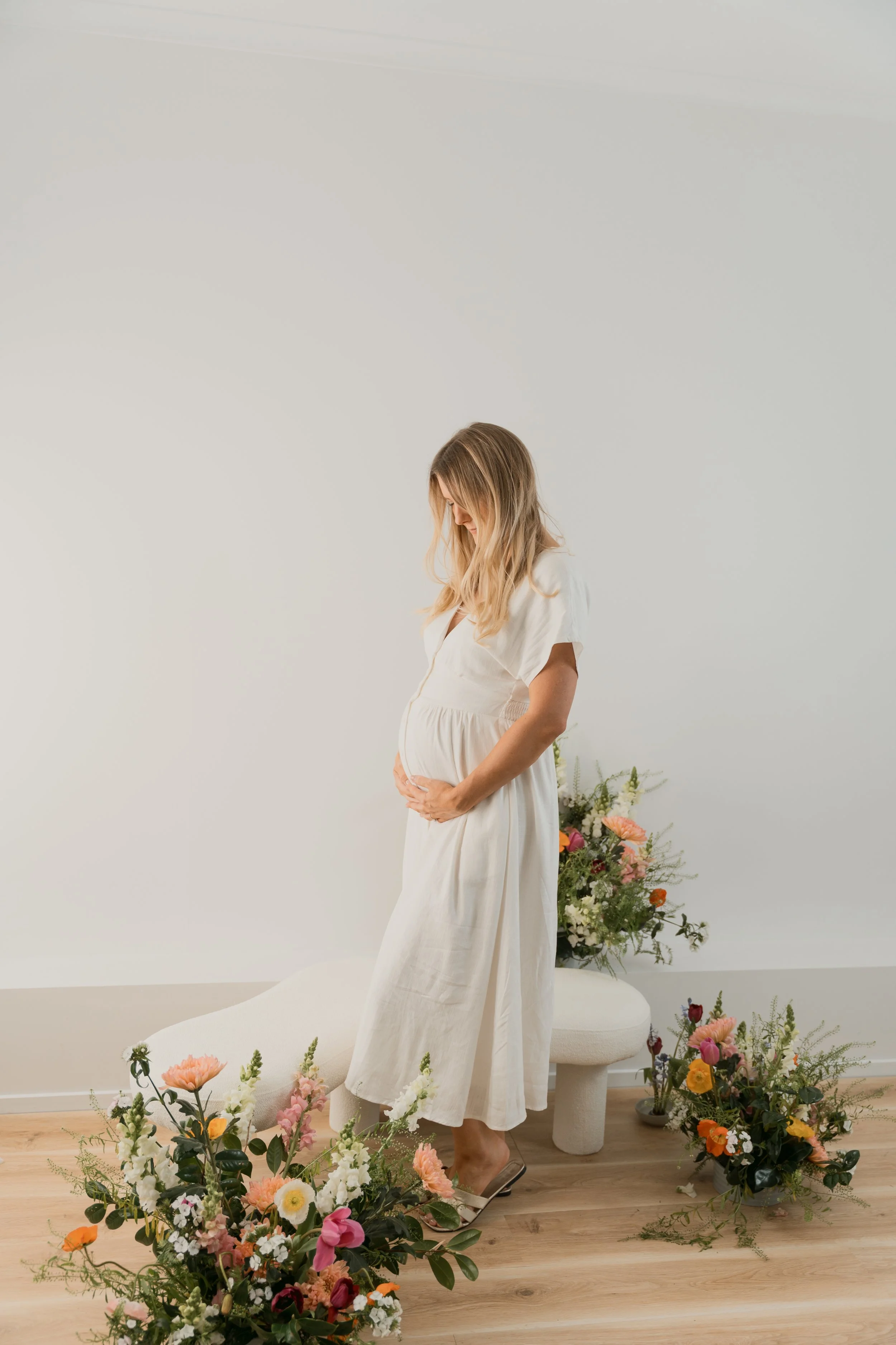 Pregnant woman in white dress standing amidst an arrangement of colorful flowers in a bright, minimalistic room.