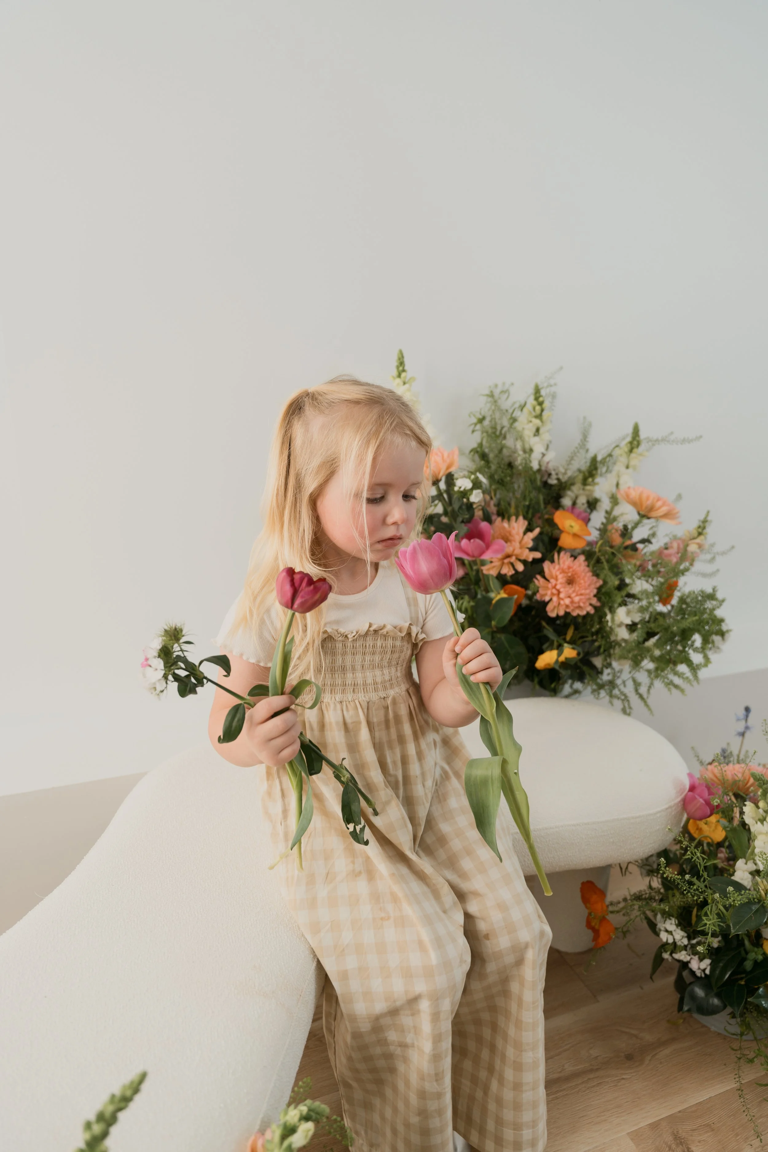 A young girl with blonde hair, wearing a beige and checkered dress, sitting in a room filled with colorful flowers, holding pink tulips.