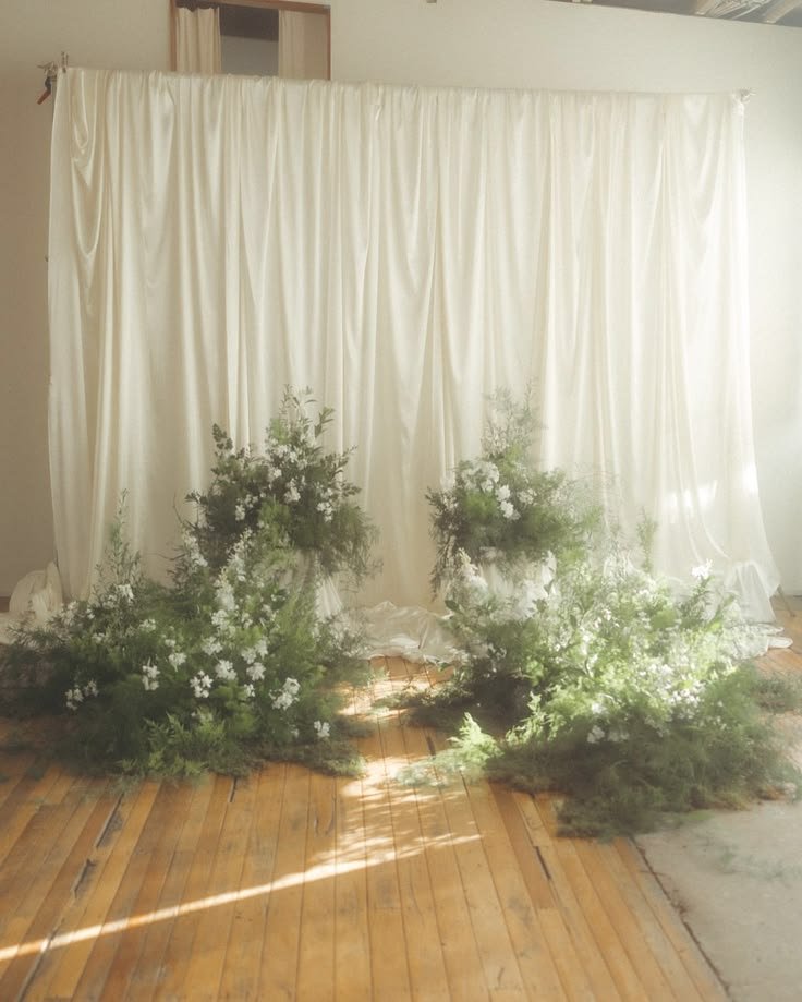 A backdrop of white curtains with green leafy plants and white flowers arranged at the base.