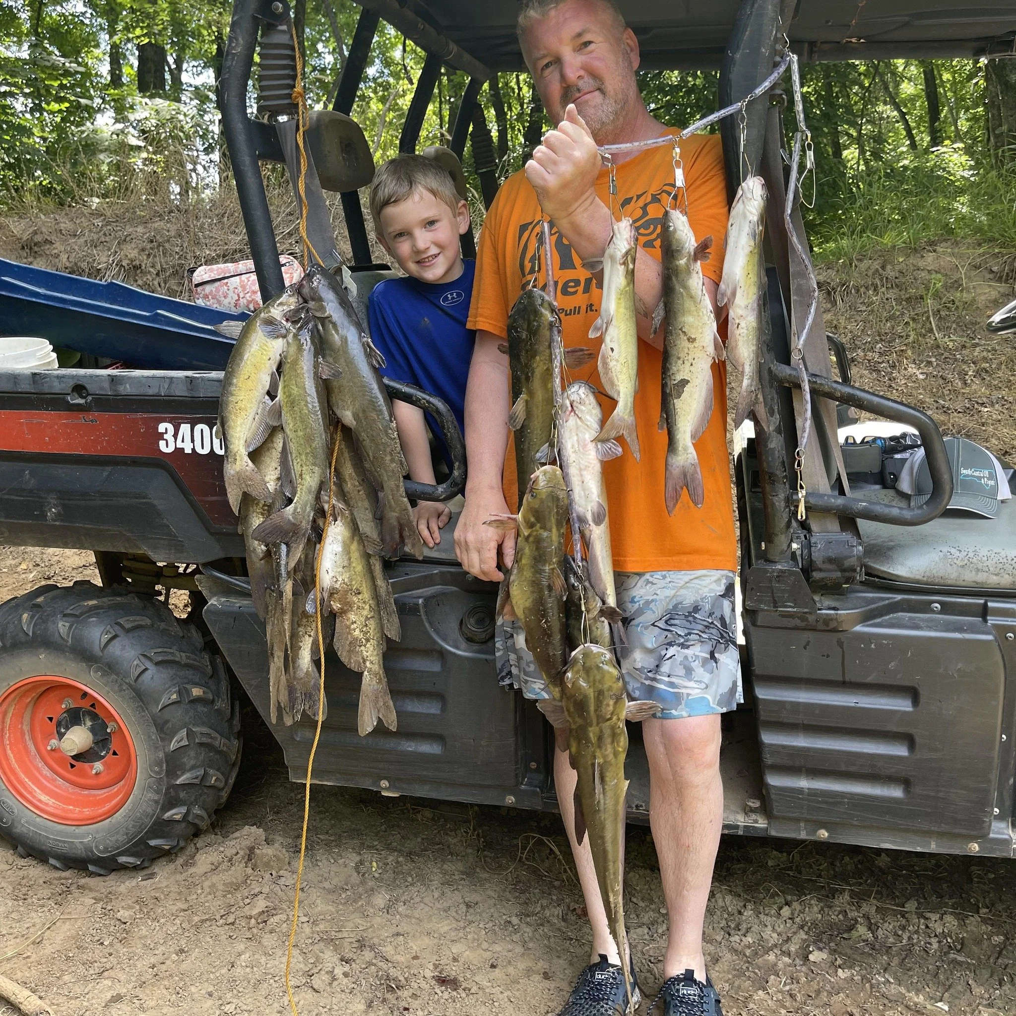 A man and a boy standing in front of a utility vehicle, holding a string of fish caught during a fishing trip, outdoors with trees in the background.