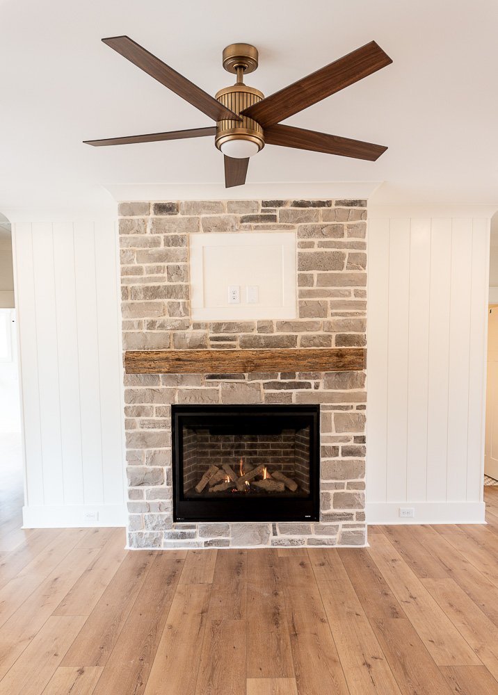 Living room with a stone fireplace, wooden mantel, white shiplap walls, hardwood floor, and ceiling fan with dark wooden blades.