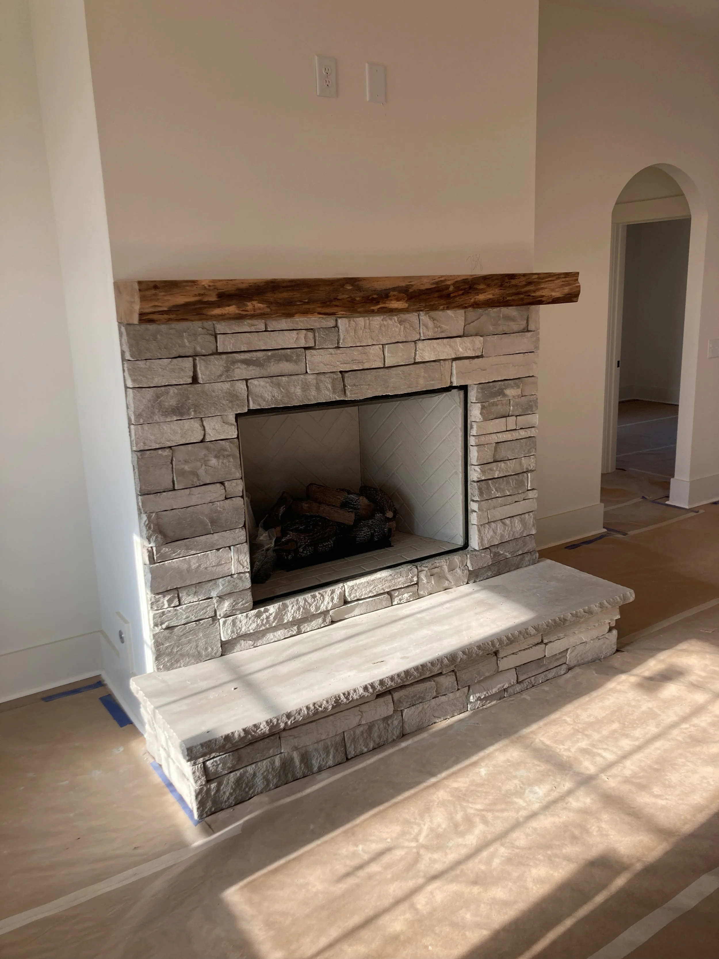 A stone fireplace with a wooden mantel in a room under construction, with protective tape on the floor and today’s sunlight.