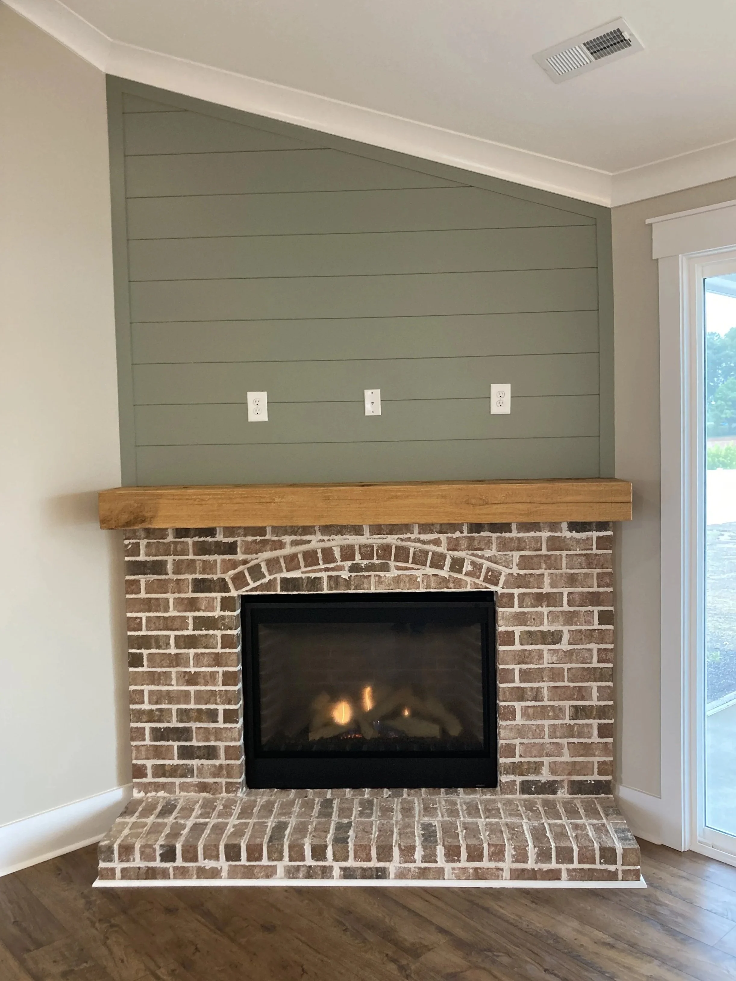 A brick fireplace with a black frame around the fire opening, a wooden mantel above, and a green shiplap wall behind it. There are three electrical outlets on the wall, and a wood floor in front of the fireplace. To the right, there is a window.