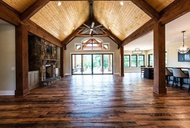 Empty living room with vaulted wooden ceiling, hardwood floors, large glass sliding door, stone fireplace, and views of a backyard with trees.