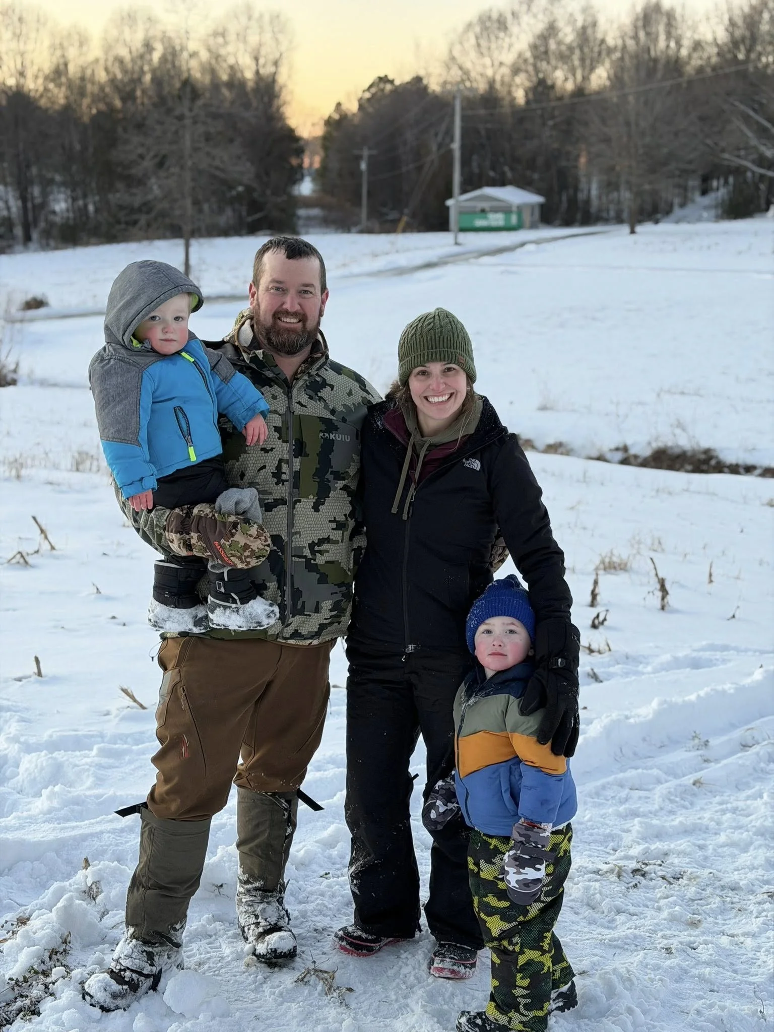 A family of five, including a man, woman, and three children, standing together in a snowy outdoor setting during winter. They are dressed in warm clothing and smiling at the camera.