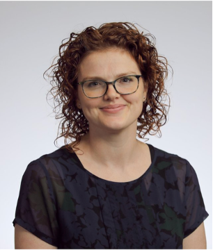 A woman with curly red hair and glasses smiling at the camera against a plain background.