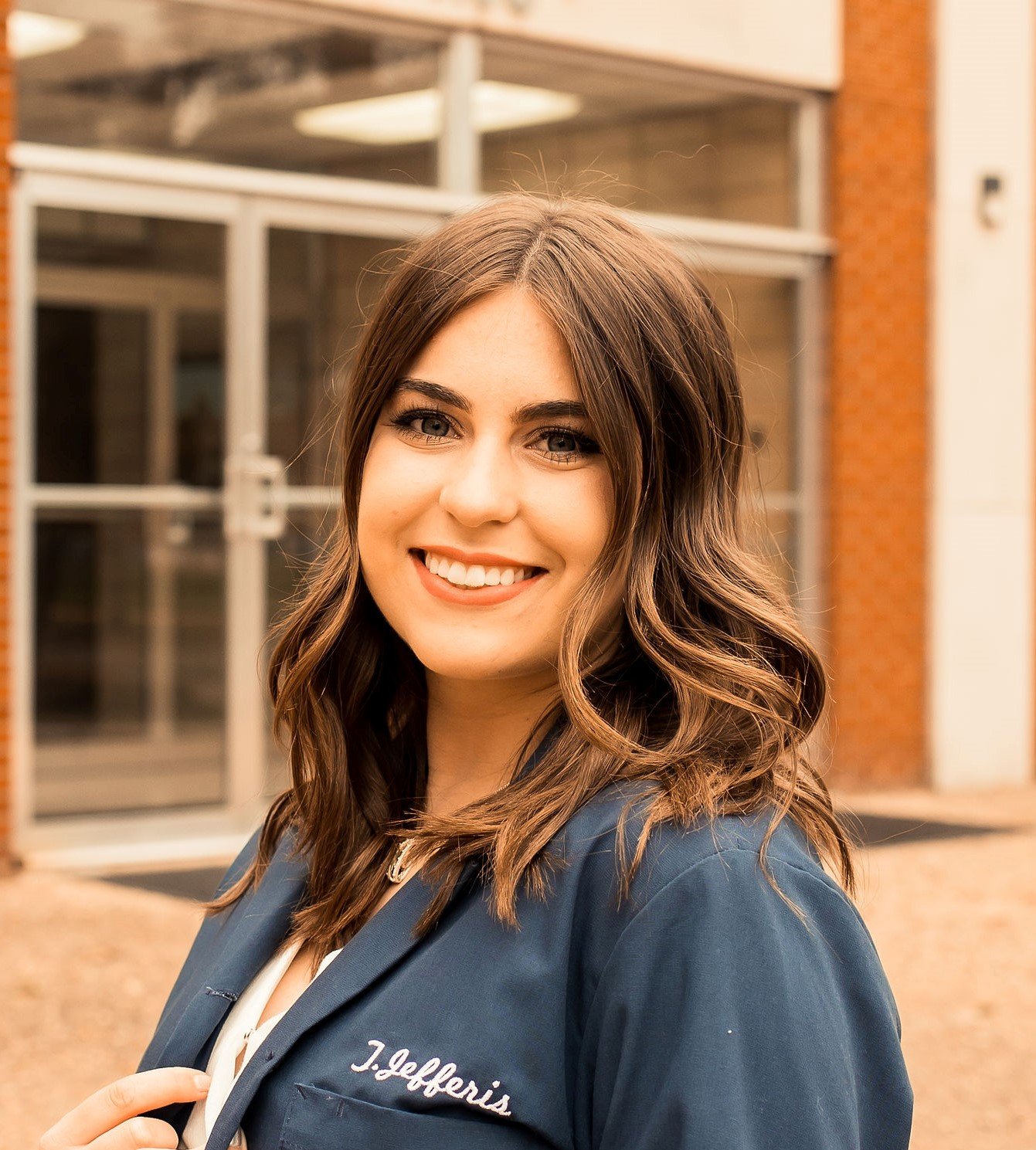 A young woman with long wavy brown hair and blue eyes, smiling, stands outside a building with glass doors and brick walls, wearing a navy blue jacket with the embroidered name 'J. Gefferis'.