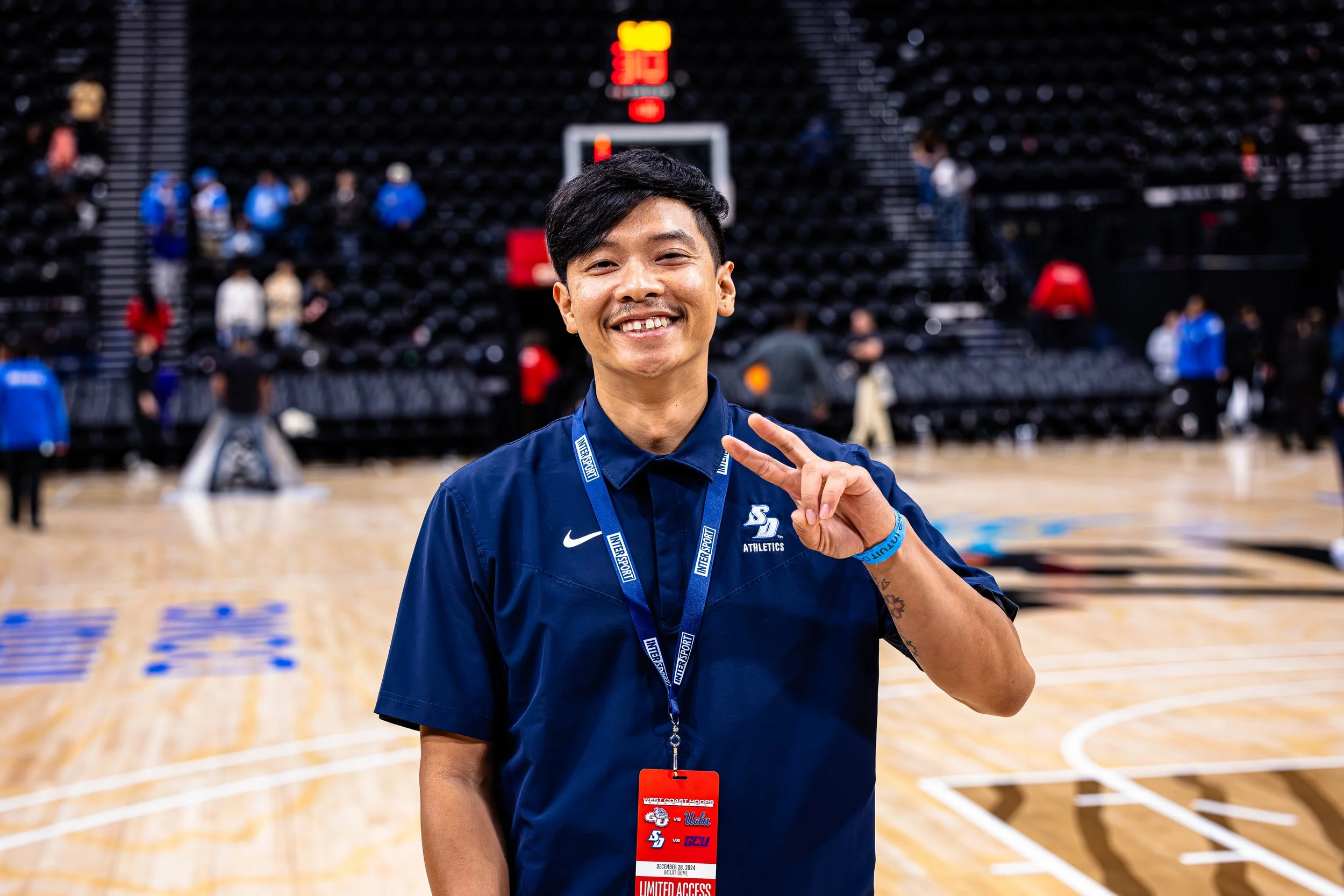 A smiling man named Justin making a peace sign at a basketball court. He is wearing a dark blue Nike shirt with the USD logo and two lanyards, one with a red pass and another blue. The background shows a gym with people and a basketball hoop.