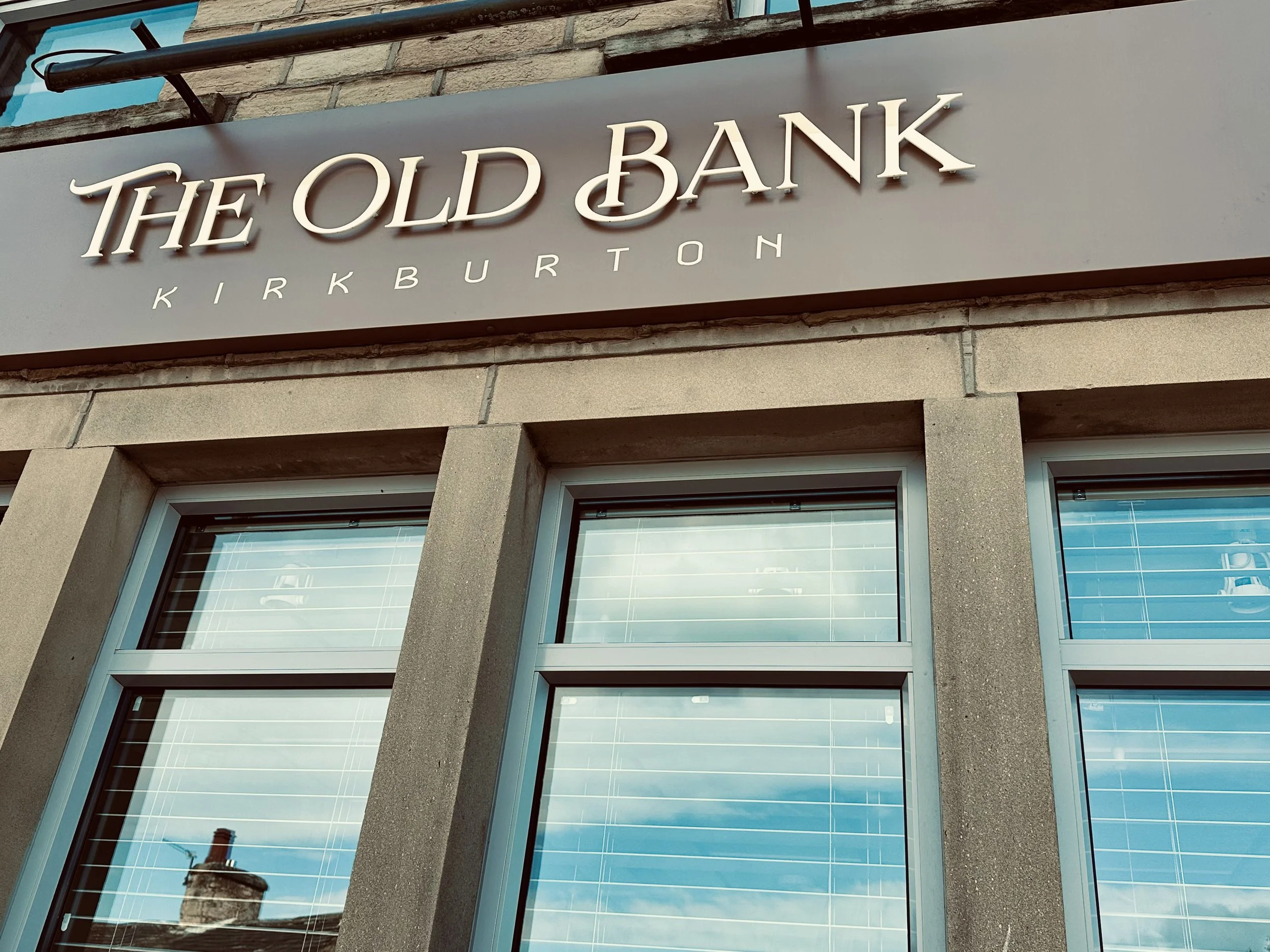 Photo of a building facade with a sign reading 'The Old Bank Kirkburton' above large windows reflecting a blue sky with clouds.
