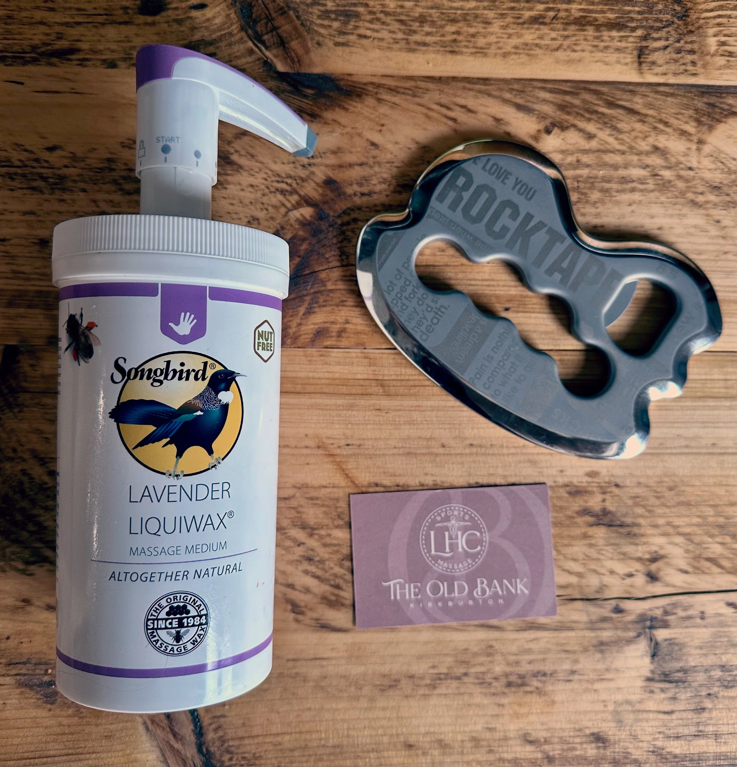 A bottle of Songbird Lavender Liquiwax massage medium, a metal bottle opener shaped like a beer bottle, and a business card for The Old Bank in Kirkcudbright on a wooden surface.