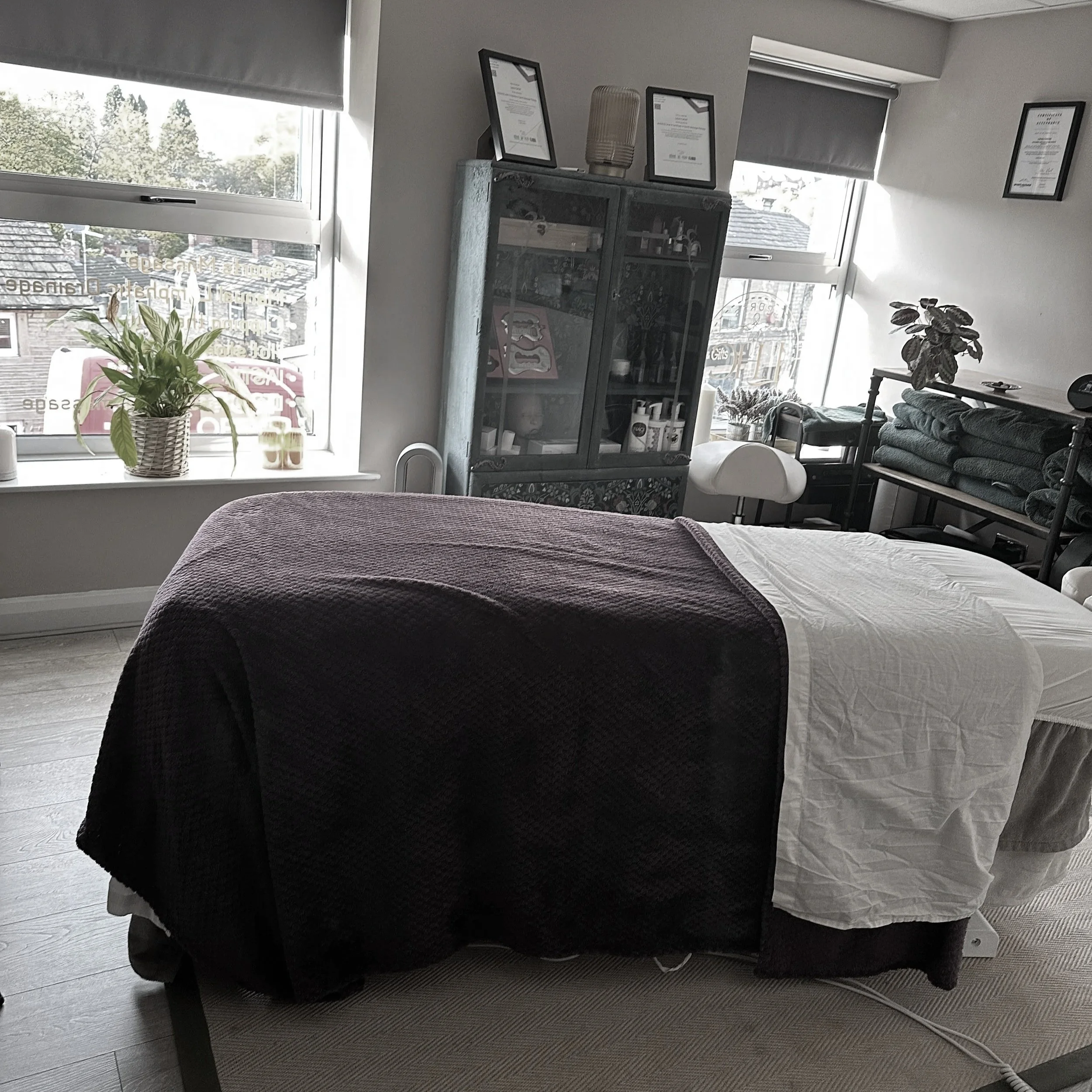 A massage table covered with a purple blanket and white sheet in a room with large windows, a cabinet with certificates and supplies, and shelves with folded towels.