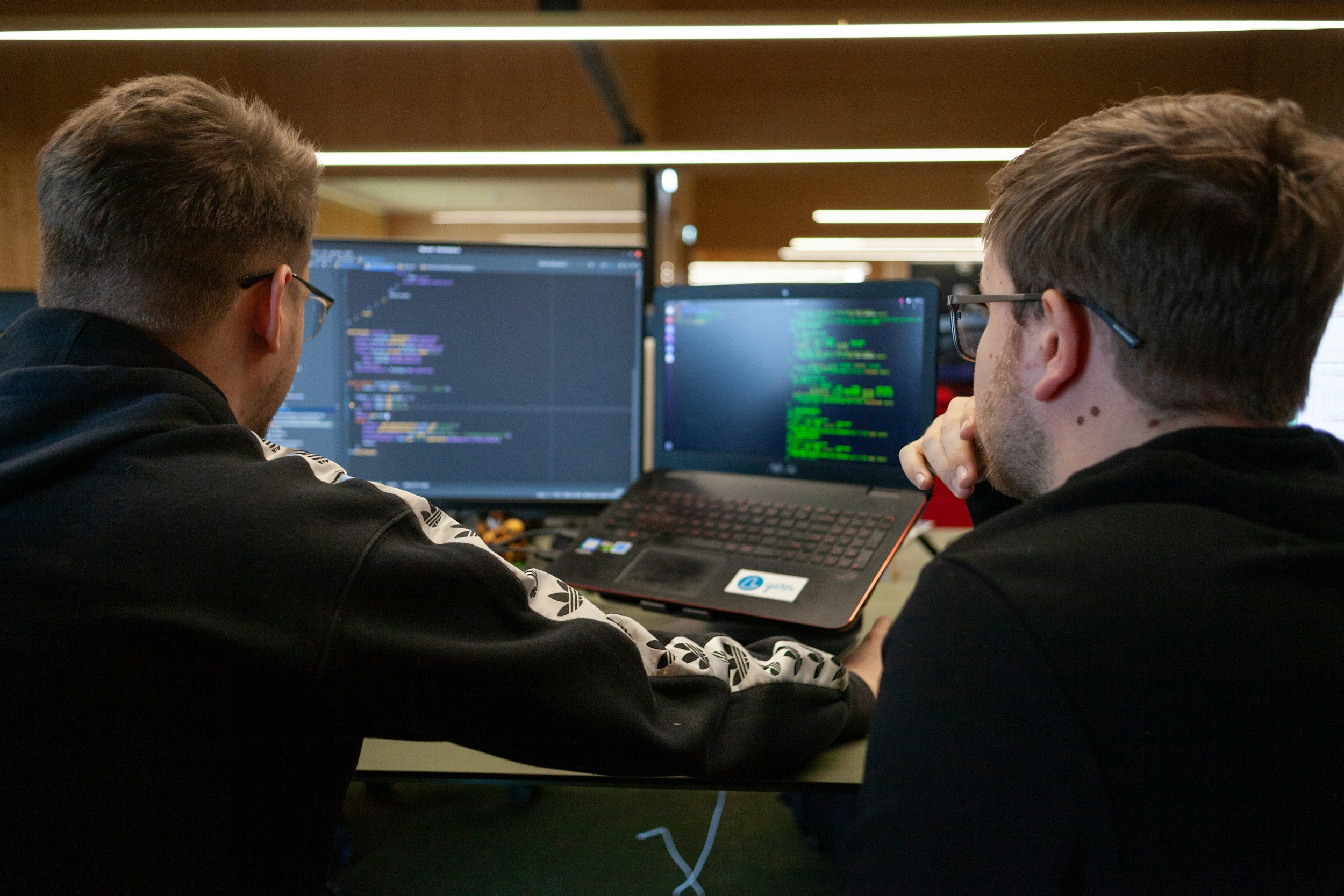 Two men working on computers with coding screens in front of them, in an office setting.