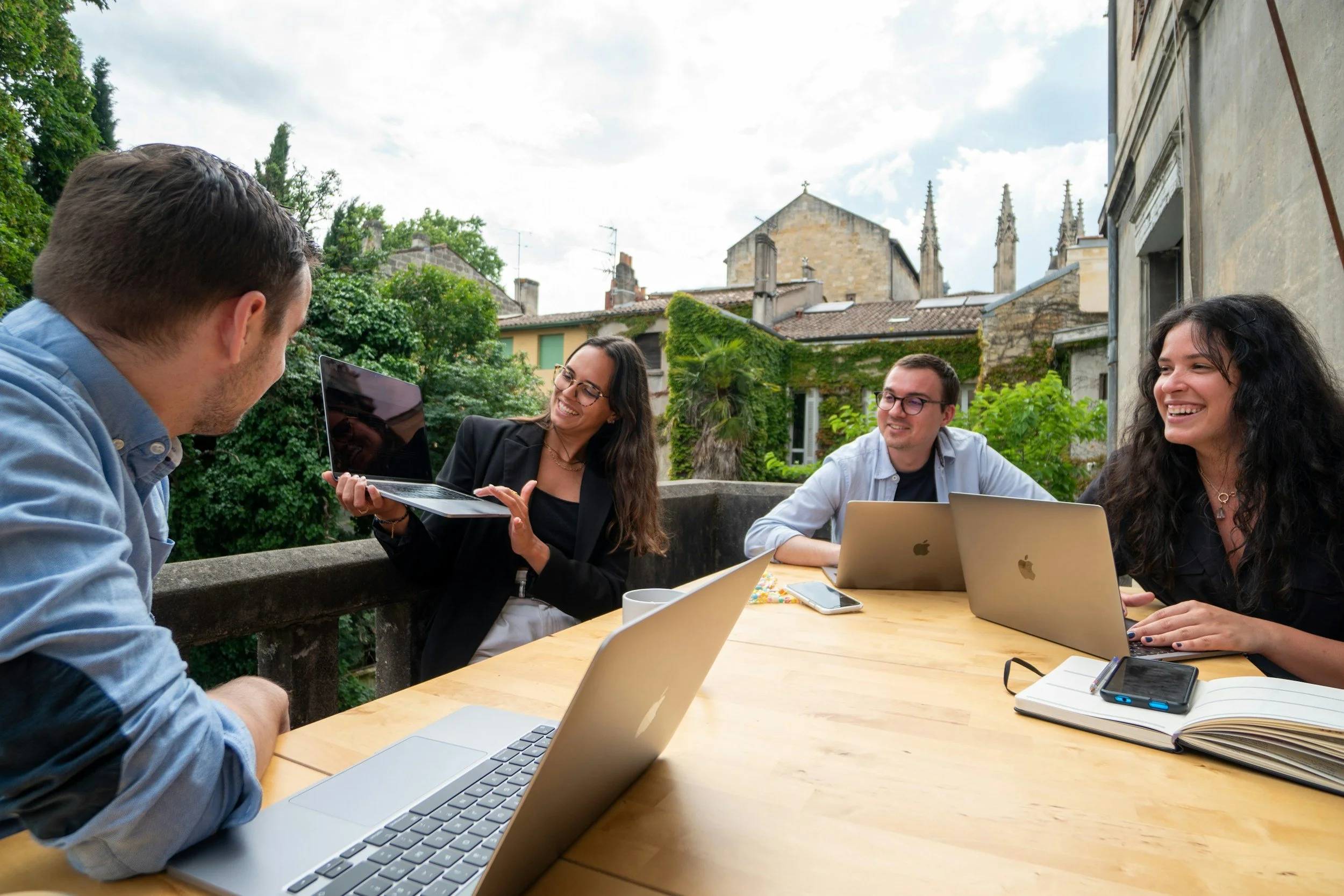 Four people sitting at a round table outdoors, engaged in conversation and smiling, with laptops, a smartphone, notebooks, and coffee mugs in front of them, with trees and historic buildings in the background.