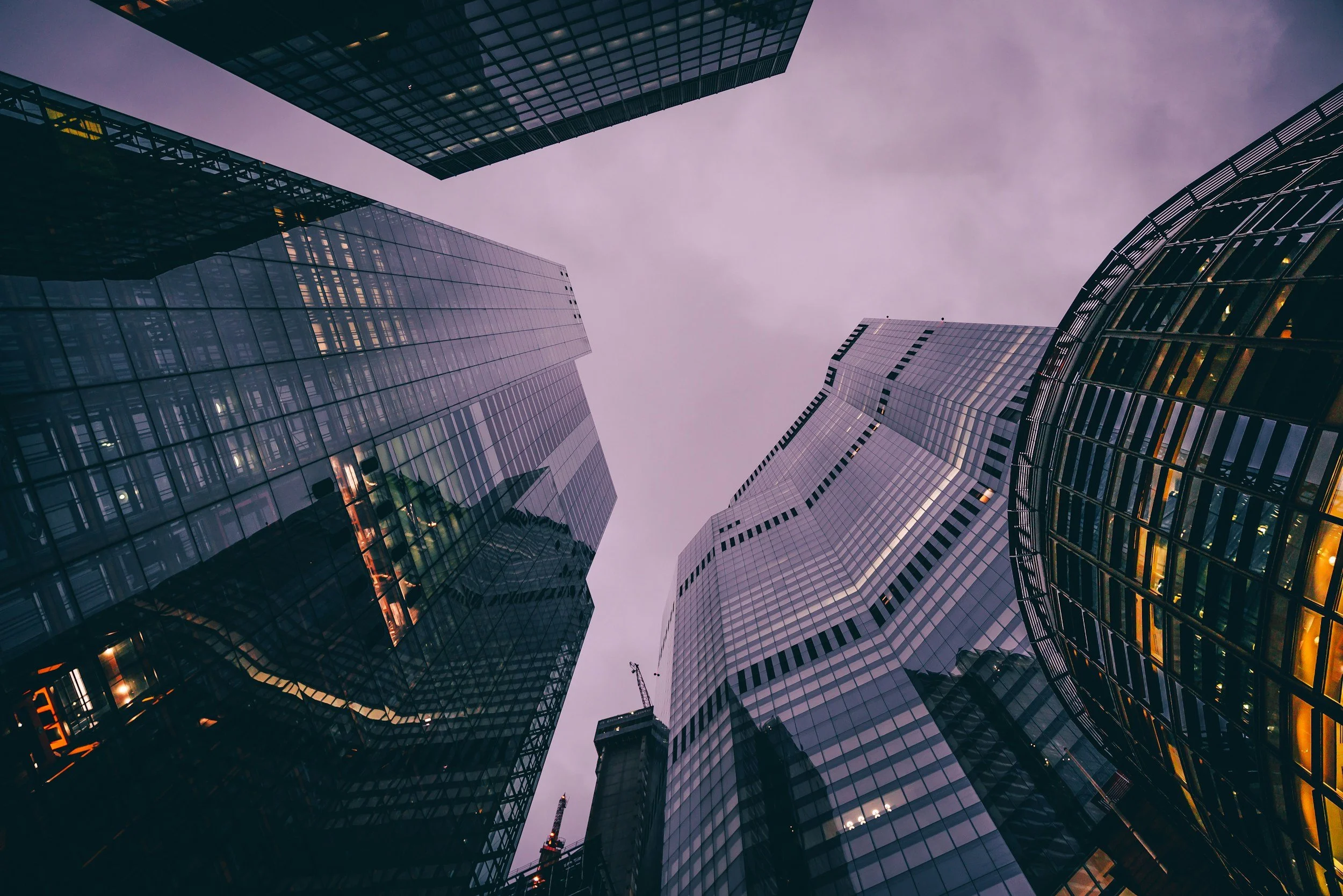 Looking up at tall glass skyscrapers in a city with illuminated windows and a cloudy purple sky.