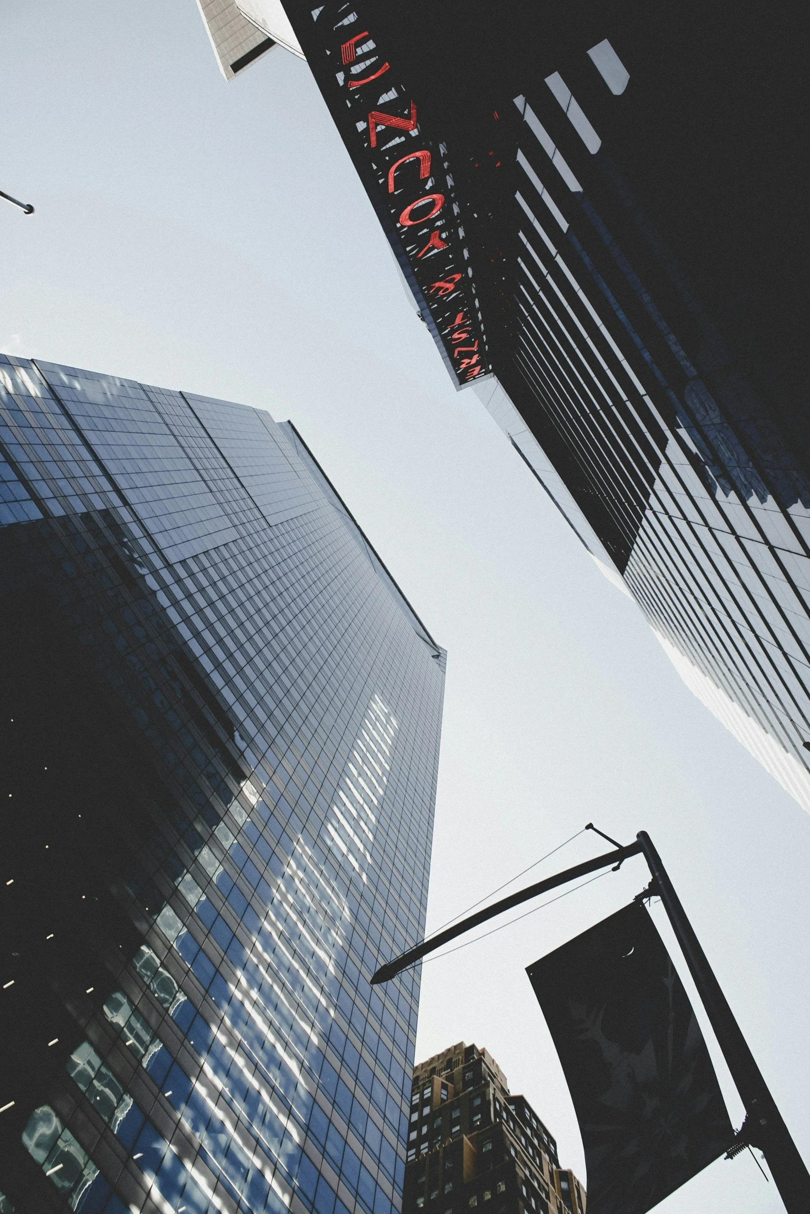View of tall glass skyscrapers and buildings in an urban city, taken from below, with a street flag and a digital sign visible in the scene.