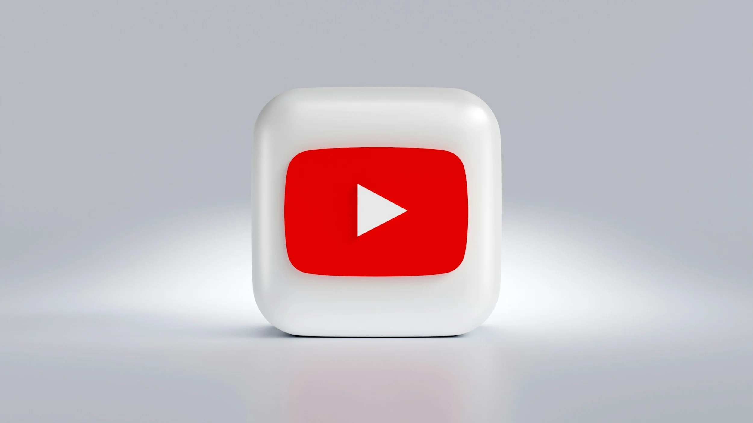 A white cube with rounded edges features the YouTube logo, which includes a red rounded rectangle with a white play button in the center, set against a plain light gray background.