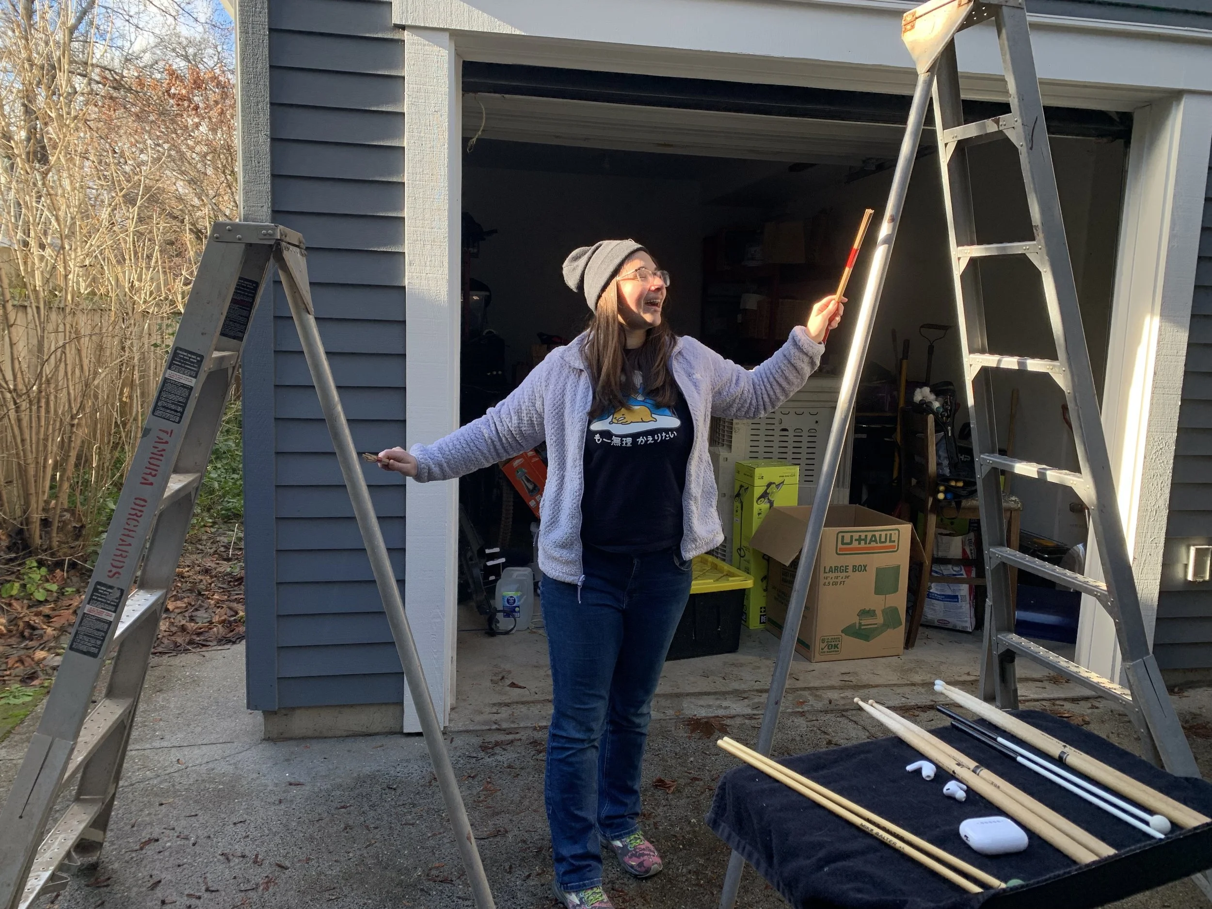 A woman standing outside a garage, smiling and holding multi rods, inbetween two ladders, with various sticks and mallets on a black music stand in front of her.