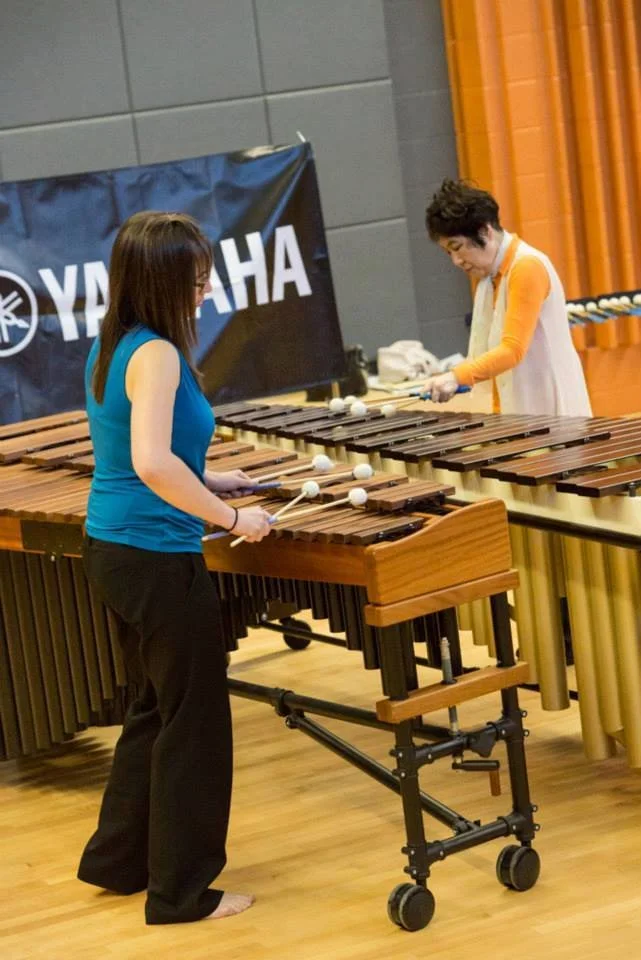 Two women, Leila Kaneda and Keiko Abe, playing marimbas in a room with gray and orange walls, and a Yamaha banner in the background.