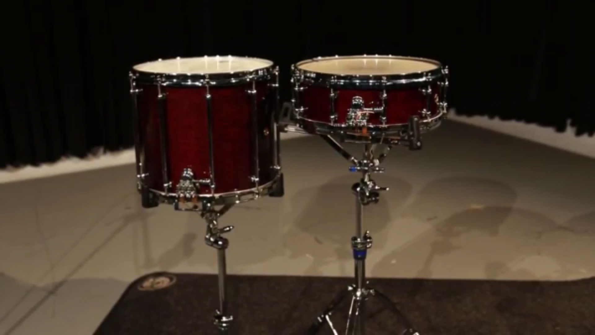 Two red drums on stands, one taller and one shorter, placed on a carpeted floor in front of a black soundproof curtain.