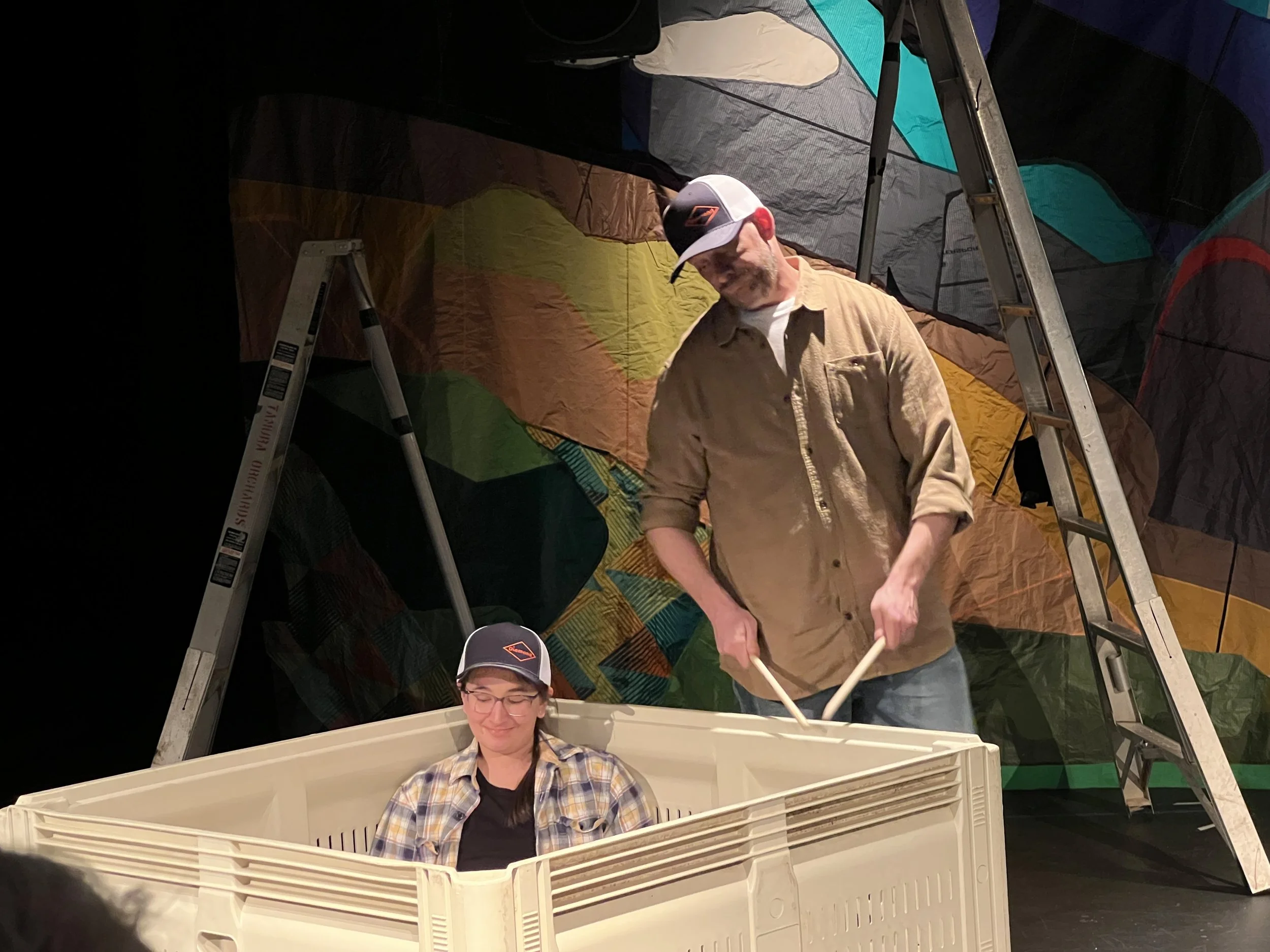 A man and a woman with hats are performing a musical act in Hood River, Oregon. The woman is in a large plastic orchard bin, and the man is drumming on the side of the bin. Ladders and a colorful tarp background are visible.