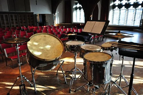 A multi-percussion setup featuring drums of various sizes, cymbals, and a music stand with sheet music inside a concert hall with rows of empty red chairs and large arched windows.