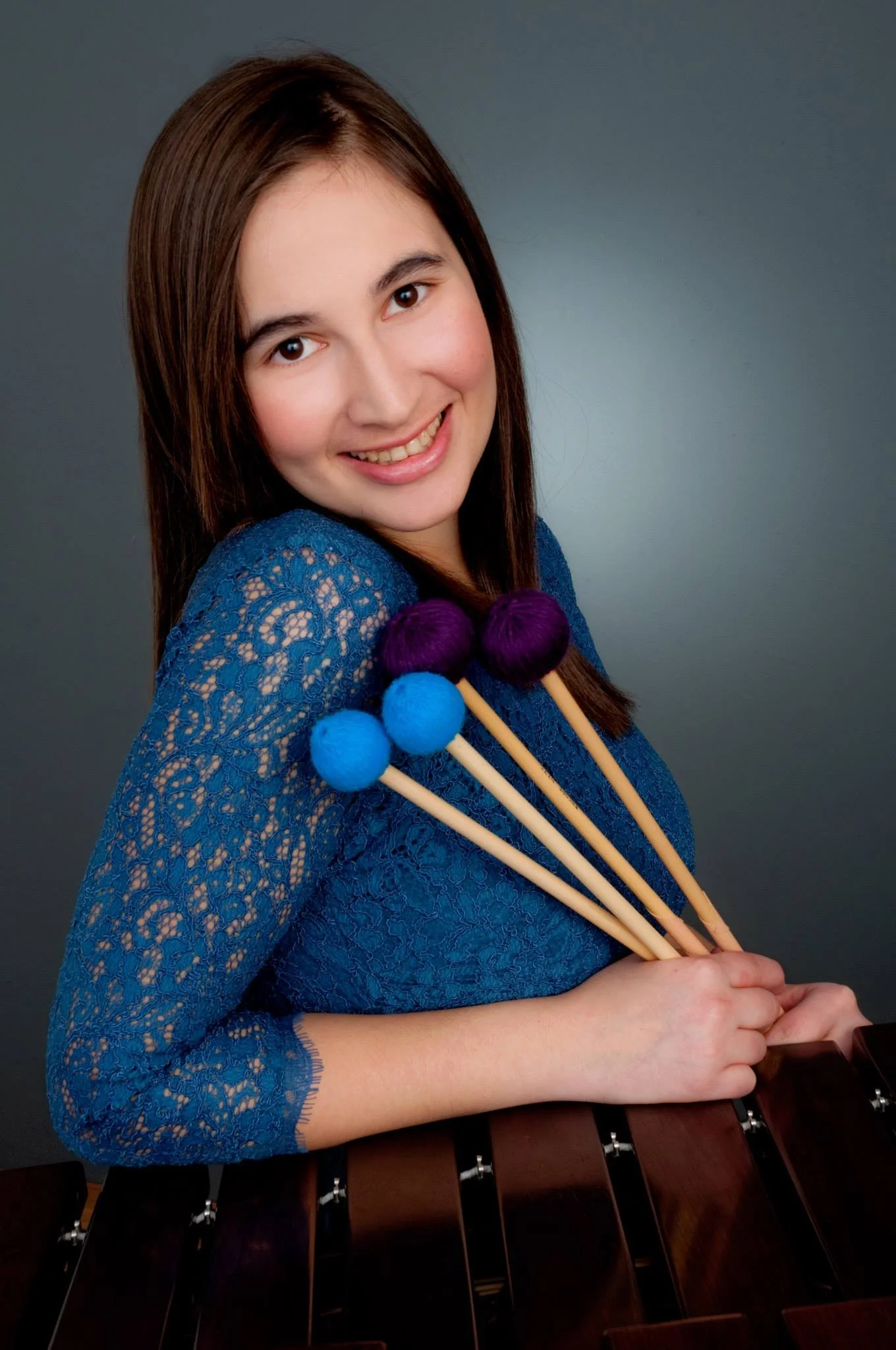 A young woman with long brown hair, smiling, wearing a blue lace dress, holding four colorful mallets with purple and blue felt heads, standing behind a marimba.