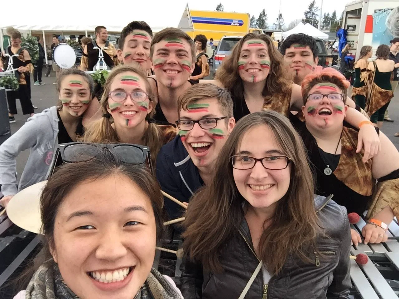 Group of young people taking a selfie at an outdoor music event, with painted faces featuring red, green, and black stripes. They are smiling and seem excited. In the background, there are more people, tents, and vehicles.