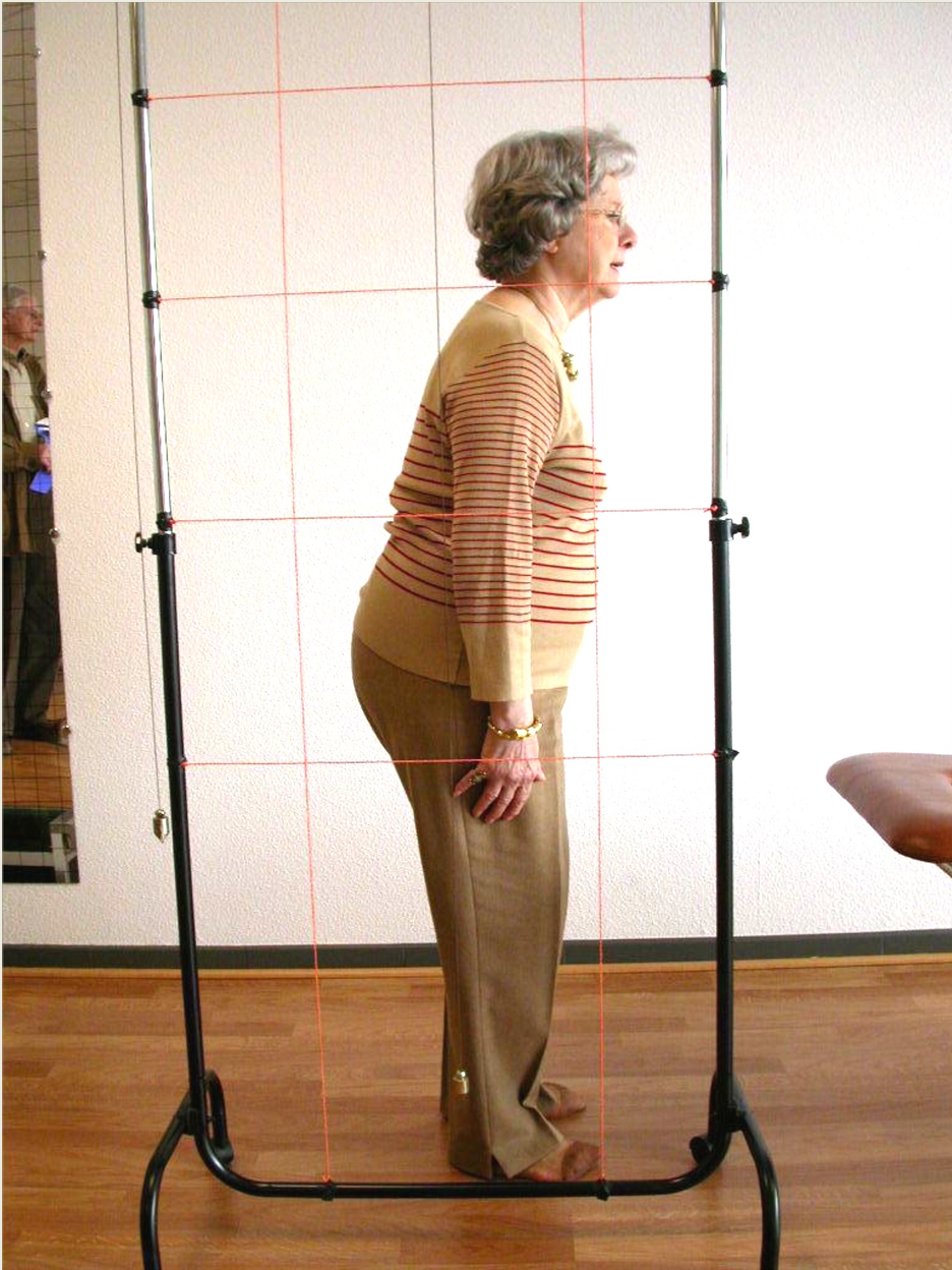An older woman with gray hair stands inside a metal grid frame with red guiding lines. She is wearing a beige sweater with red stripes, beige pants, and a gold bracelet and necklace. She appears to be in an indoor setting with a light-colored textured wall and wooden floor. There is a mirror on the left reflecting part of the room and another person partially visible.