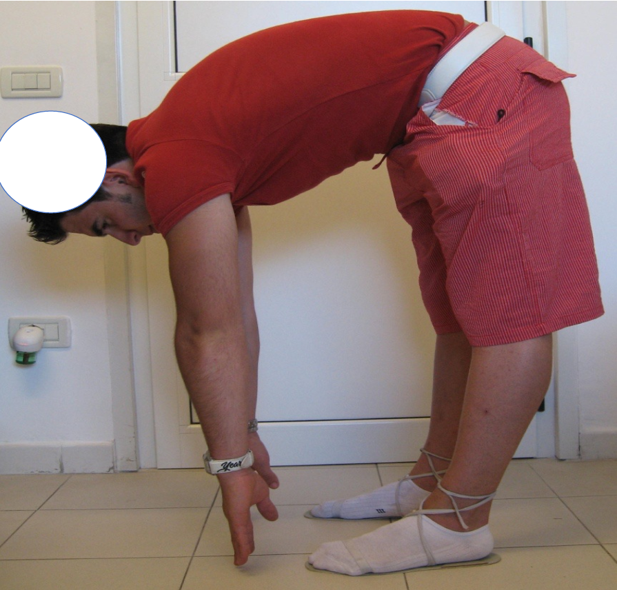 Man bending forward in yoga pose, hands and feet on the floor, wearing a red shirt and red striped shorts, indoors with a white wall and door in the background.