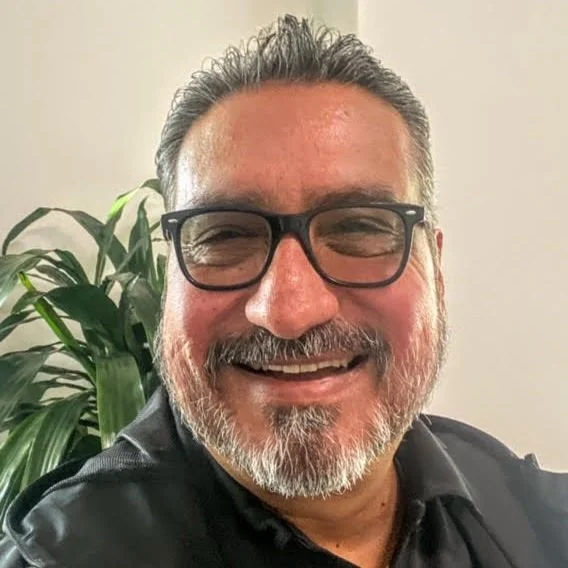 Smiling man with glasses and a beard, sitting in front of a large green plant and a light-colored wall.