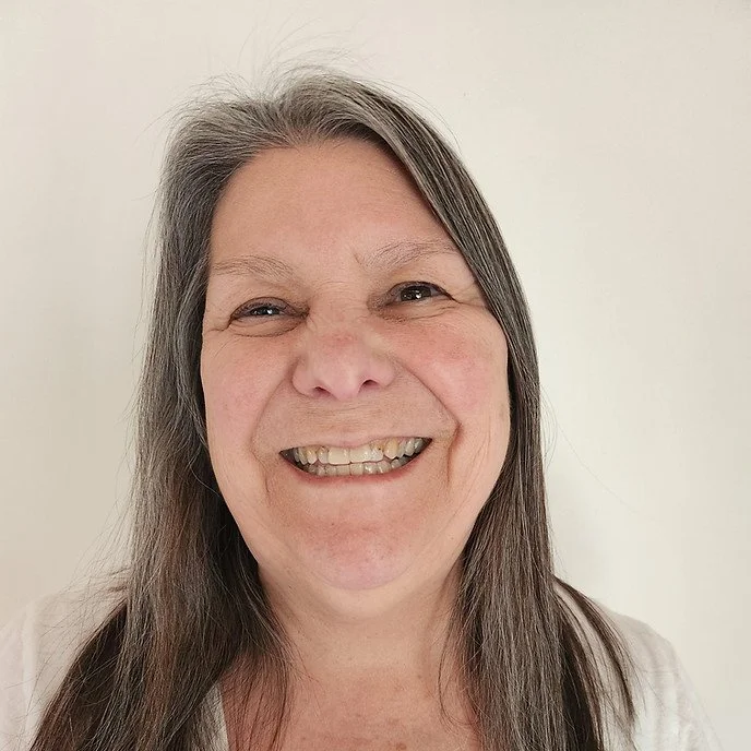 A smiling woman with long gray hair and light skin, standing against a plain light-colored background.