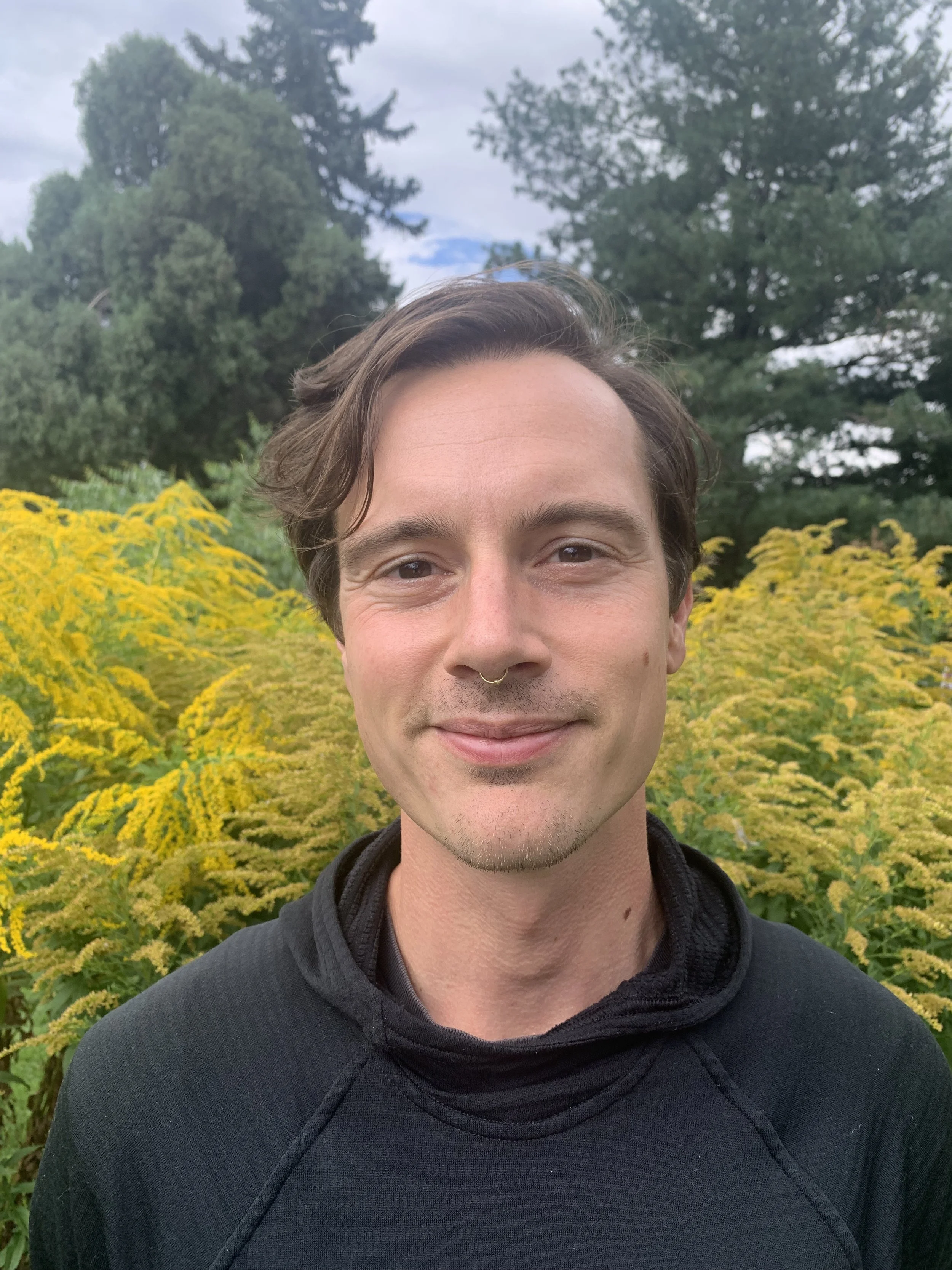 A person with short brown hair, wearing a black shirt, smiling outdoors in front of yellow flowers and green trees under a cloudy sky.
