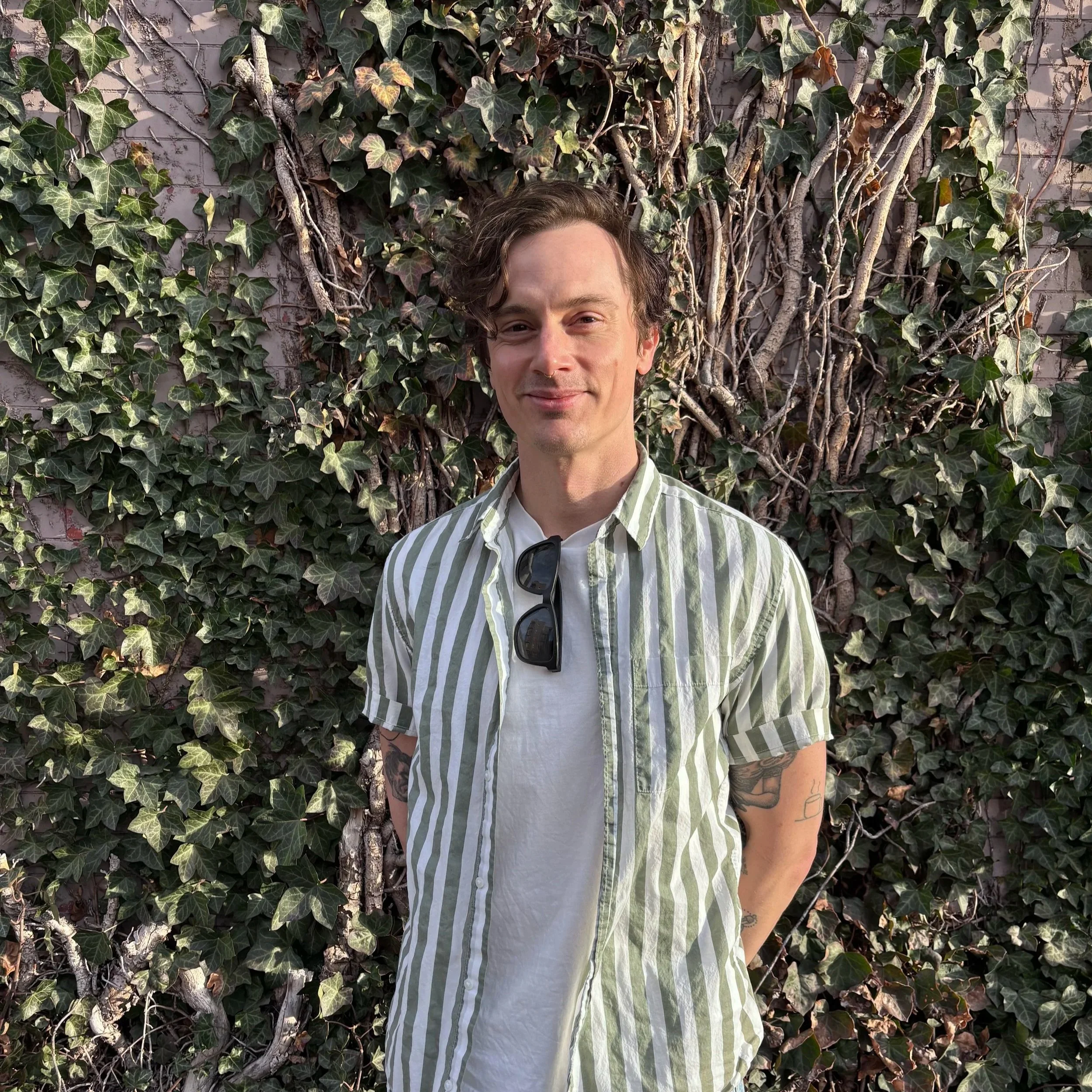 A man with brown, wavy hair stands in front of a wall covered with green ivy, wearing a striped green and white shirt with sunglasses hanging from the collar.