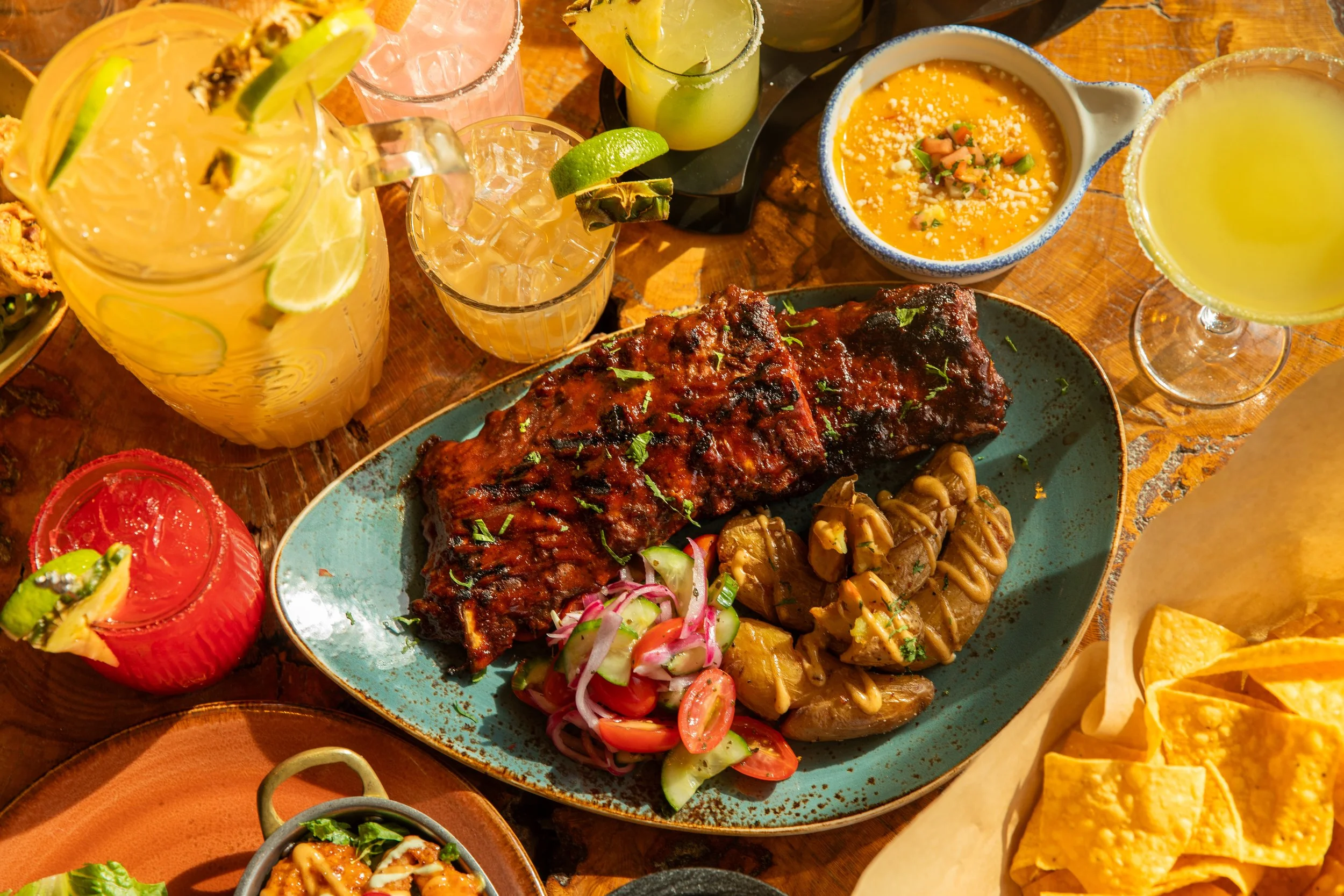Plated rack of ribs with roasted potatoes and salad, surrounded by margaritas, chips, and small bowls on a wooden table.