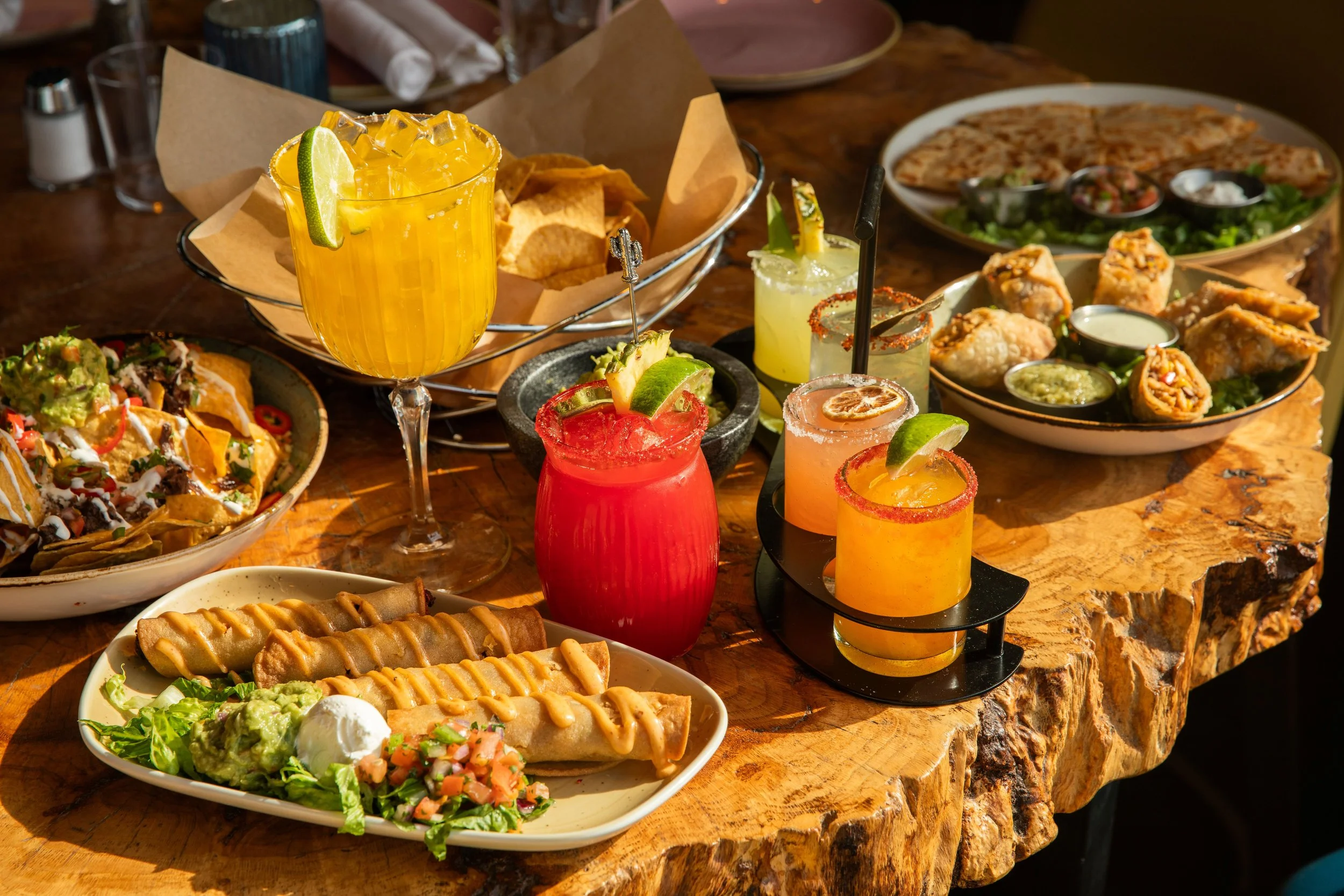Tabletop spread of folded tacos, chips and dip, and colorful margaritas and cocktails arranged on a rustic wooden table.
