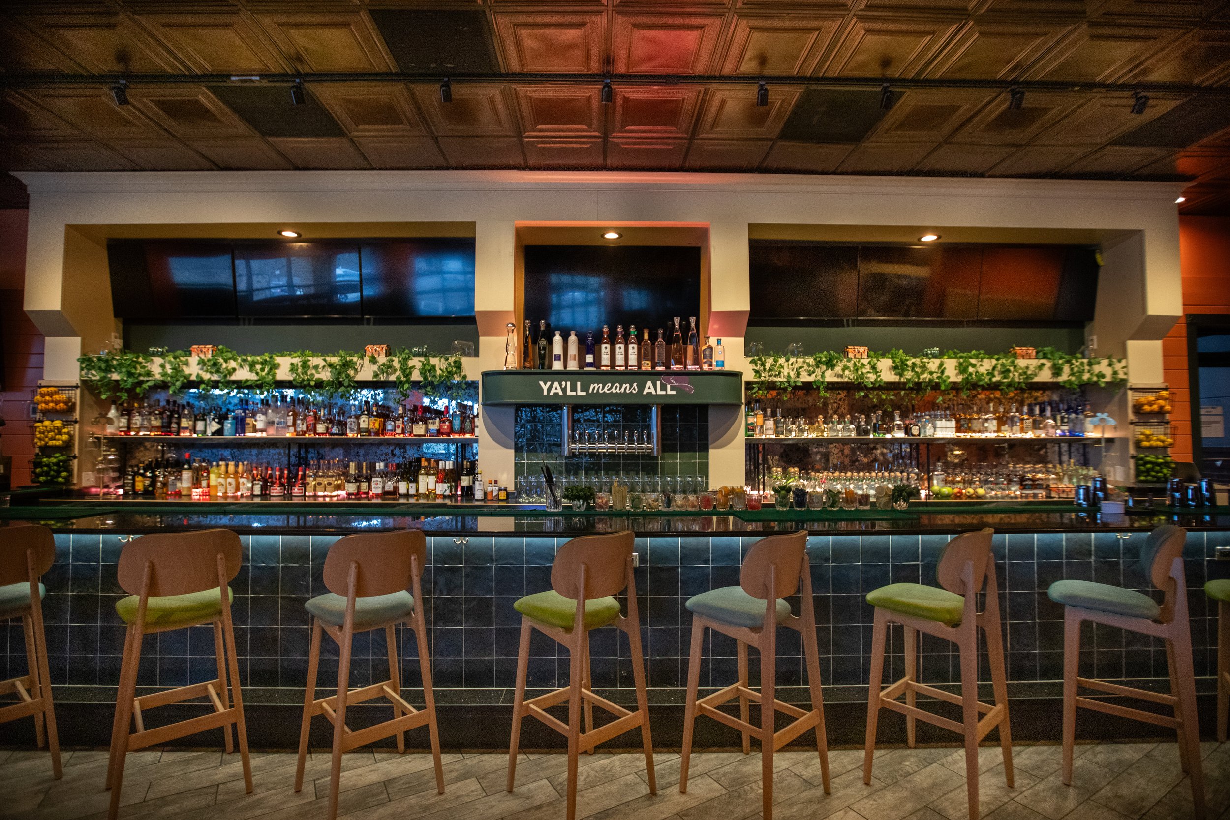 Bar with wooden stools, shelves stocked with liquor bottles and glassware, greenery accents, and a sign reading “Y’ALL means ALL” above the center.