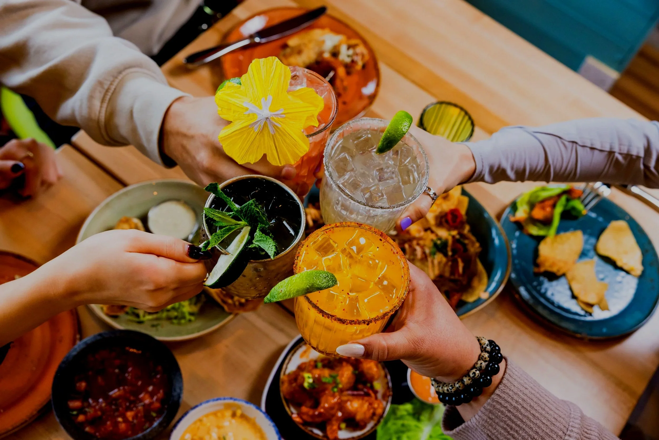 Group raising colorful cocktails over a table of shared Mexican dishes, seen from above.