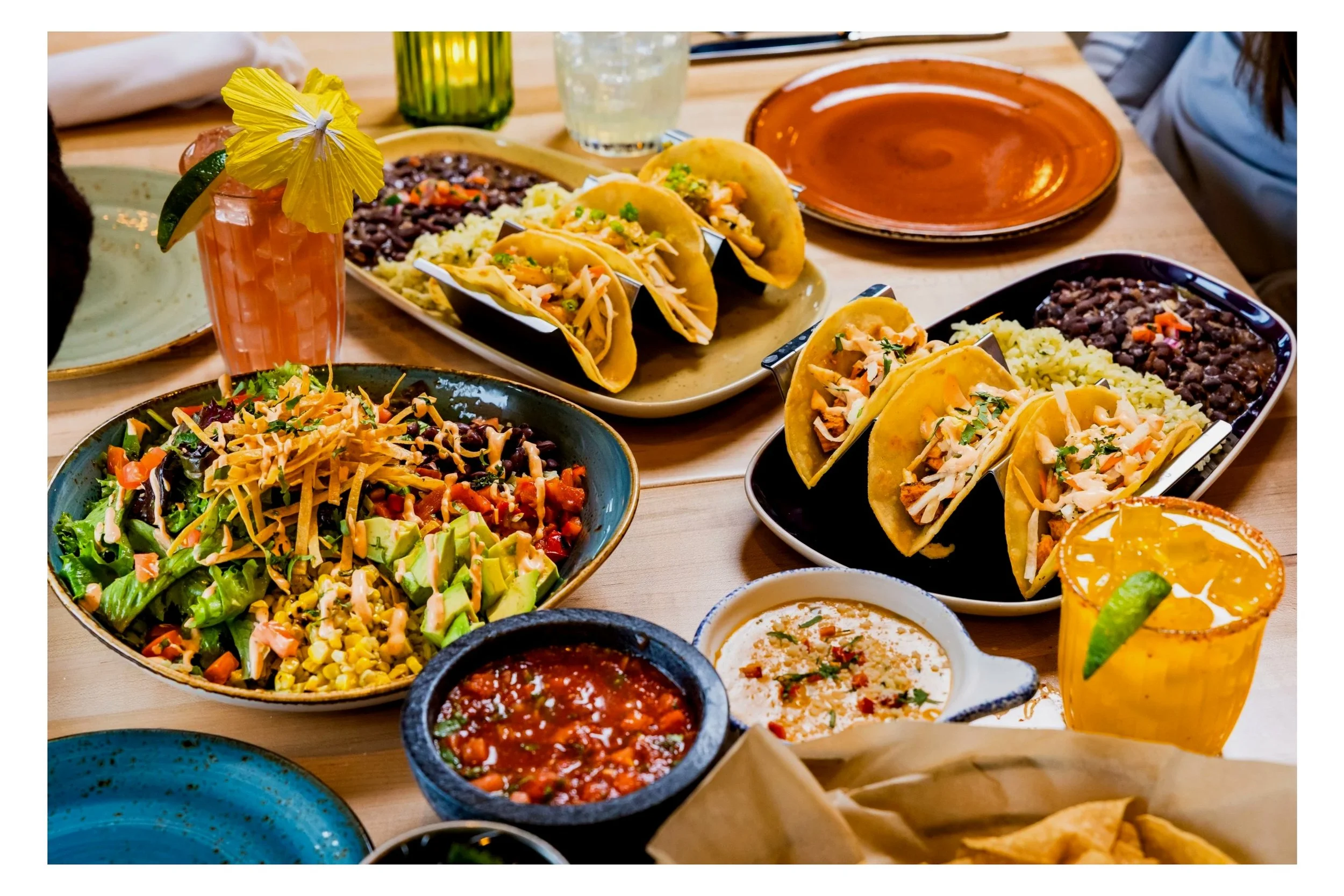 Table filled with tacos, salads, dips, and colorful drinks, arranged on assorted plates and bowls.
