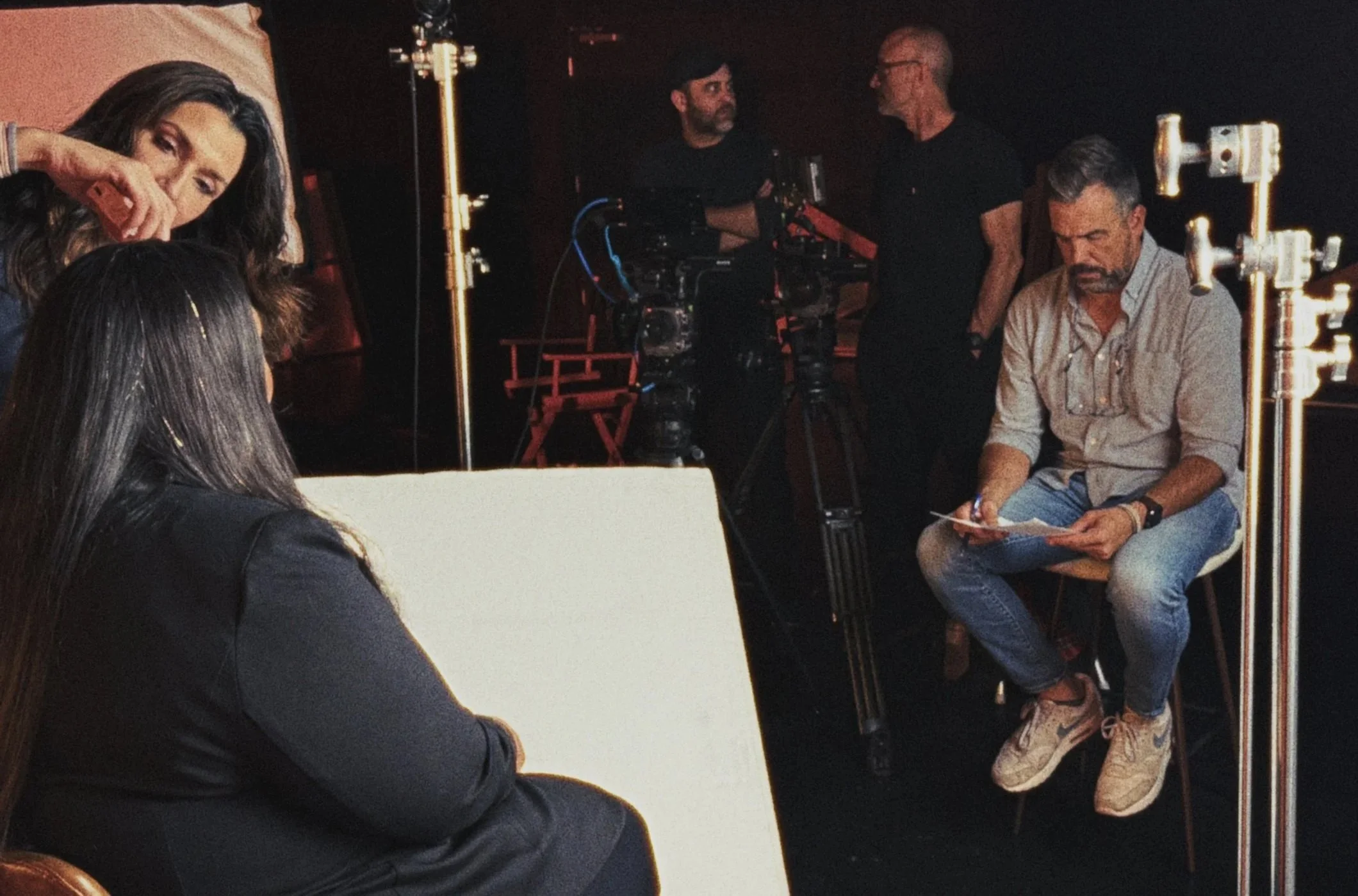 Filming crew setting up a scene with a woman in black sitting on a chair, a woman with long dark hair adjusting her hair, a man with a beard reviewing notes, and crew members with cameras and equipment in a dark studio.