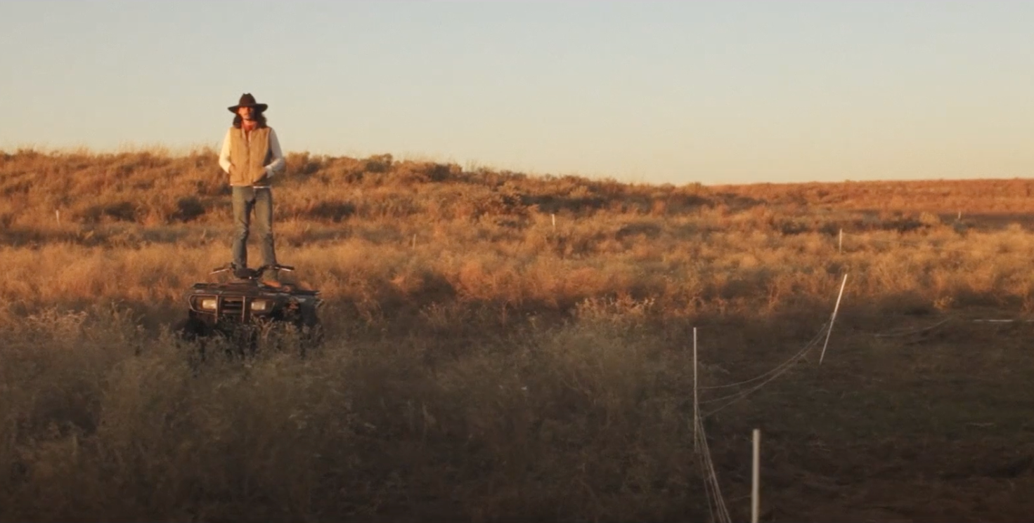 Person standing on an ATV in a desert landscape at sunset, wearing sunglasses, a wide-brimmed hat, and layered clothing.