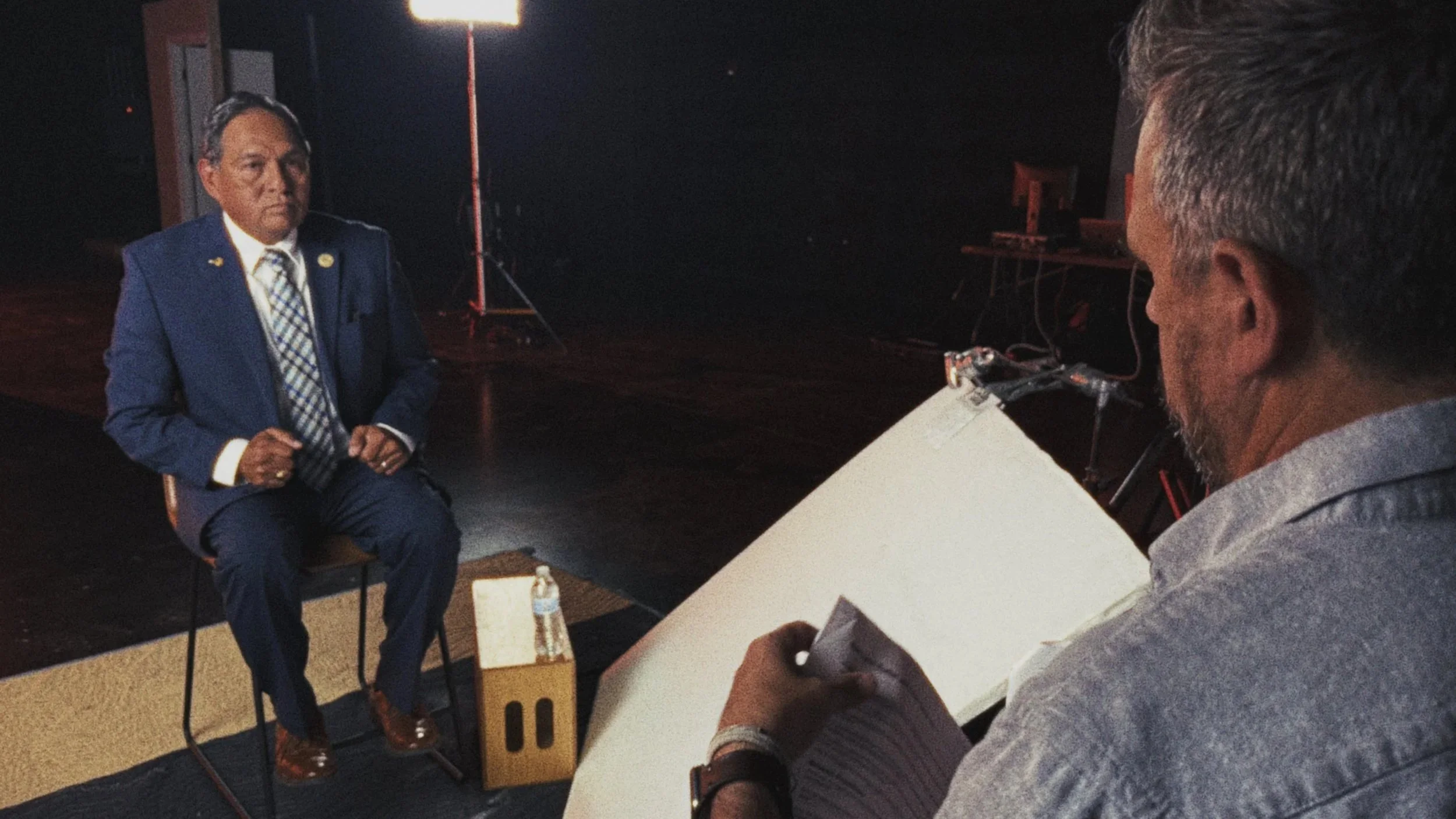 A man in a blue suit being interviewed, sitting on a chair in a studio setting with dark background, while the interviewer writes notes on a spiral notebook, with lighting equipment visible behind him.