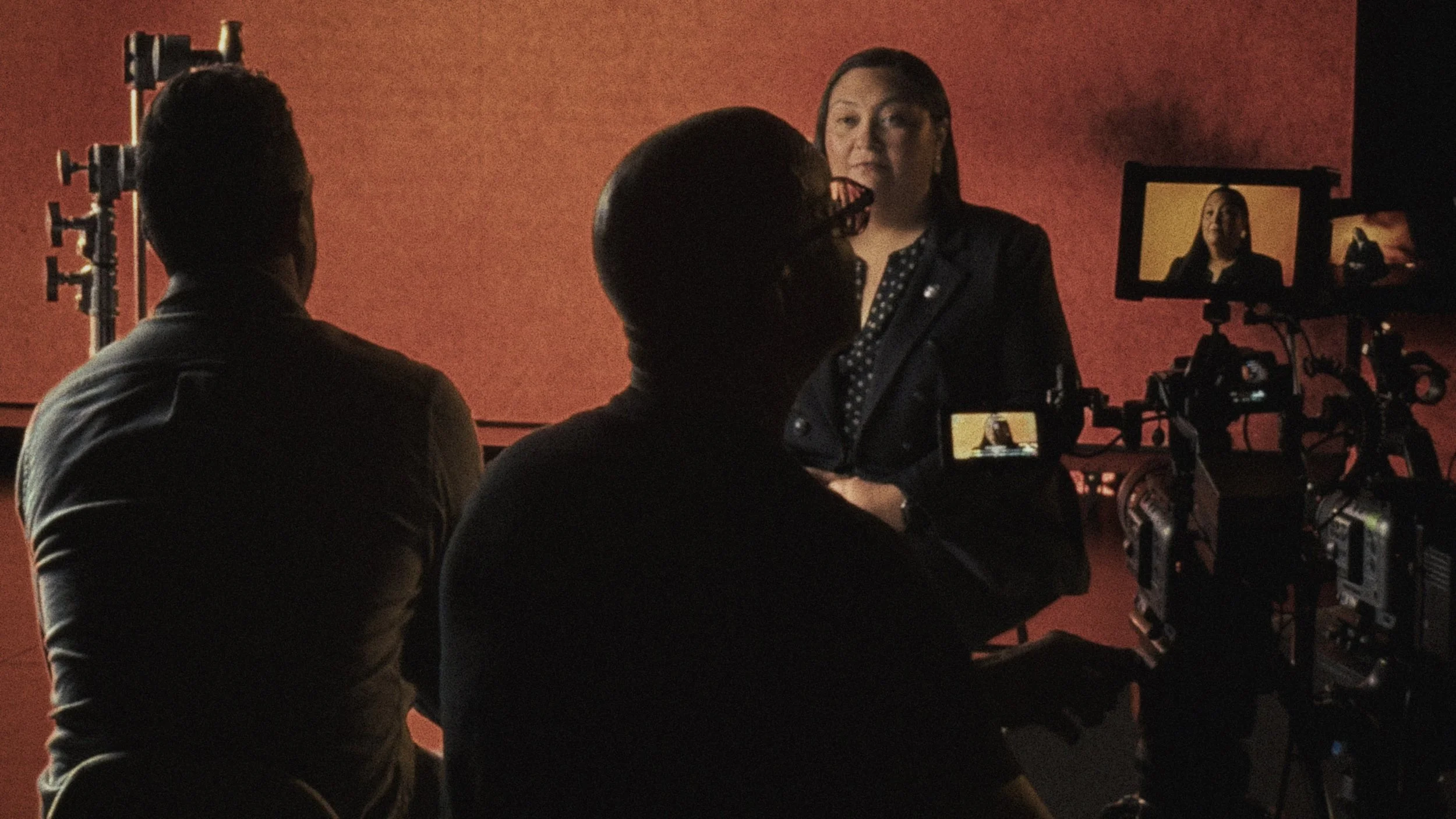 A woman being filmed during an interview, with two men operating cameras, in a studio with a red background.