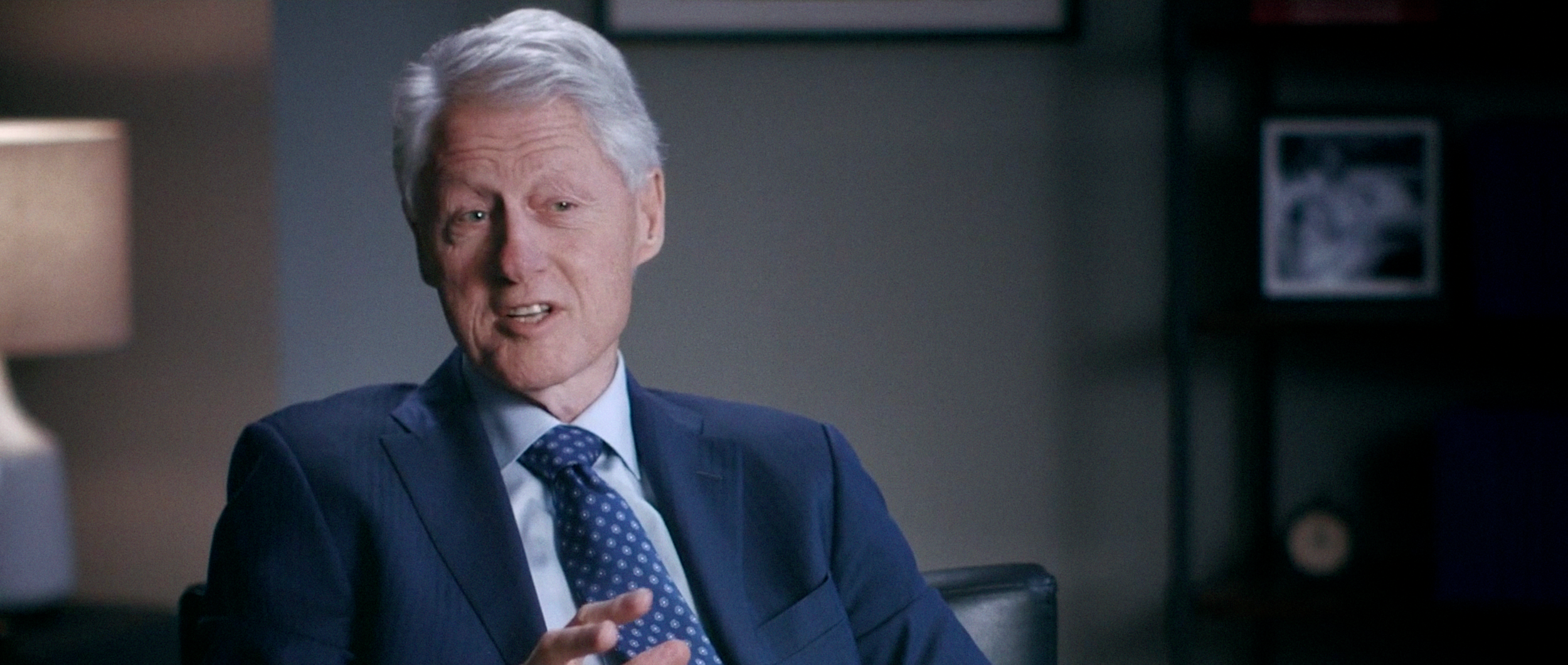A mature man with white hair in a business suit and a blue polka dot tie, sitting in an office, speaking and gesturing with his right hand.