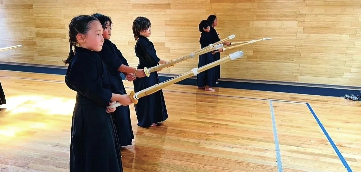 A group of young girls practicing martial arts with wooden sticks in a gymnasium, standing in a line along a wooden wall.