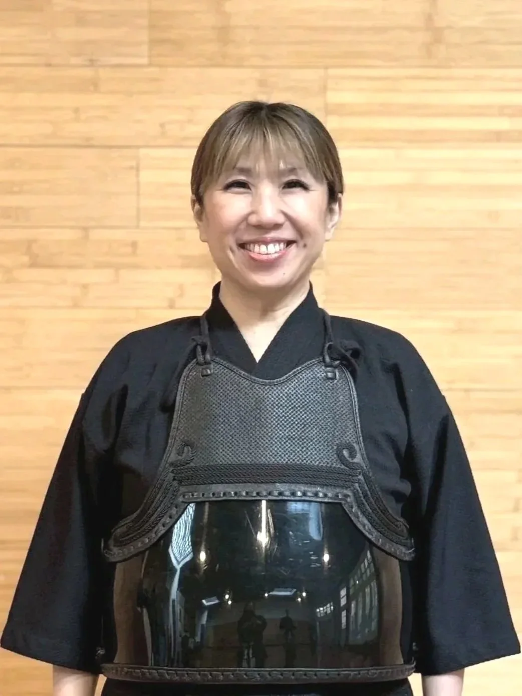 A woman with short brown hair wearing a black protectve vest over a black traditional martial arts uniform standing in front of a wooden wall, smiling at the camera.