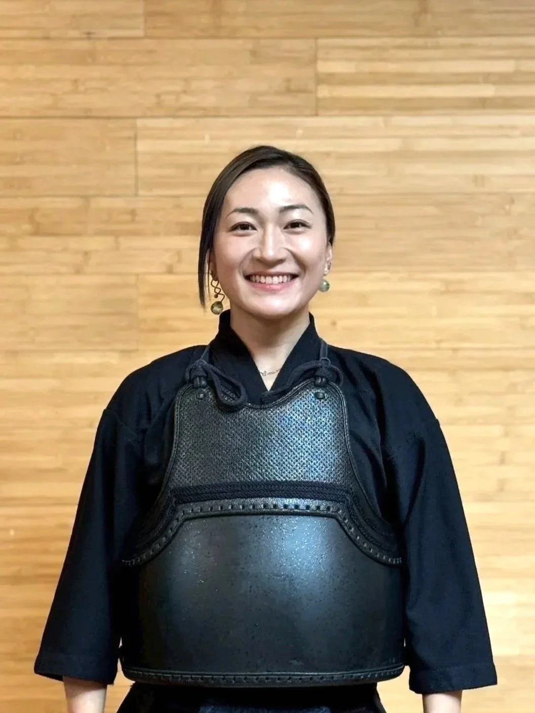 Woman in black martial arts uniform smiling, standing in front of a wooden wall, wearing a black chest protector and earrings.