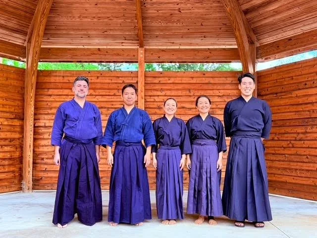 Five people dressed in traditional martial arts uniform standing in a semi-open wooden pavilion, smiling at the camera.