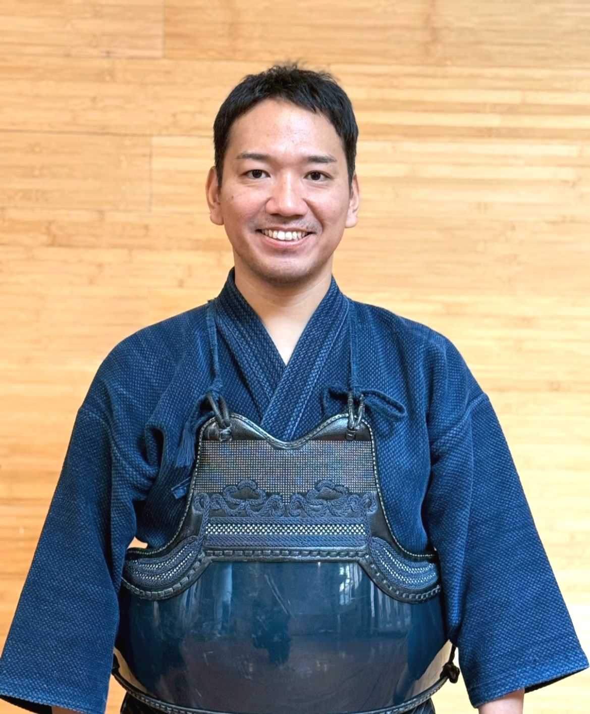 Man in traditional martial arts uniform wearing protective gear, standing indoors in front of a wooden wall.