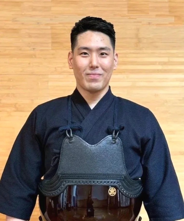 Young man in black martial arts uniform wearing a chest protector against a wooden wall.