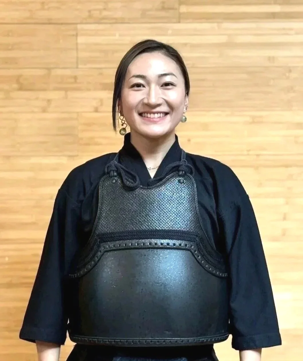Woman in black martial arts uniform smiling, standing in front of a wooden wall, wearing a black chest protector and earrings.